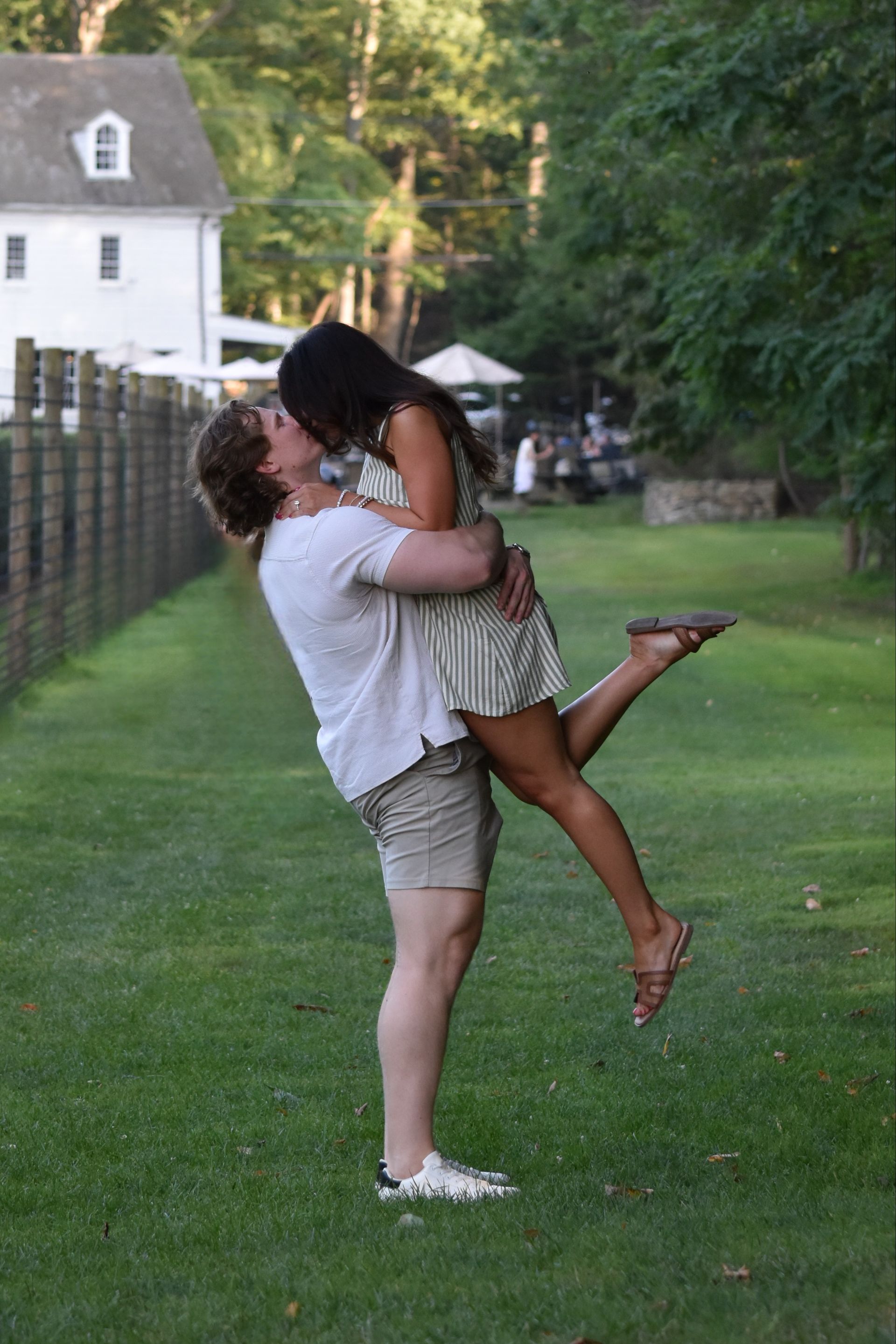 Couple embraces outdoors. Man lifts woman, kissing. Green grass, trees, and a white building in the background.