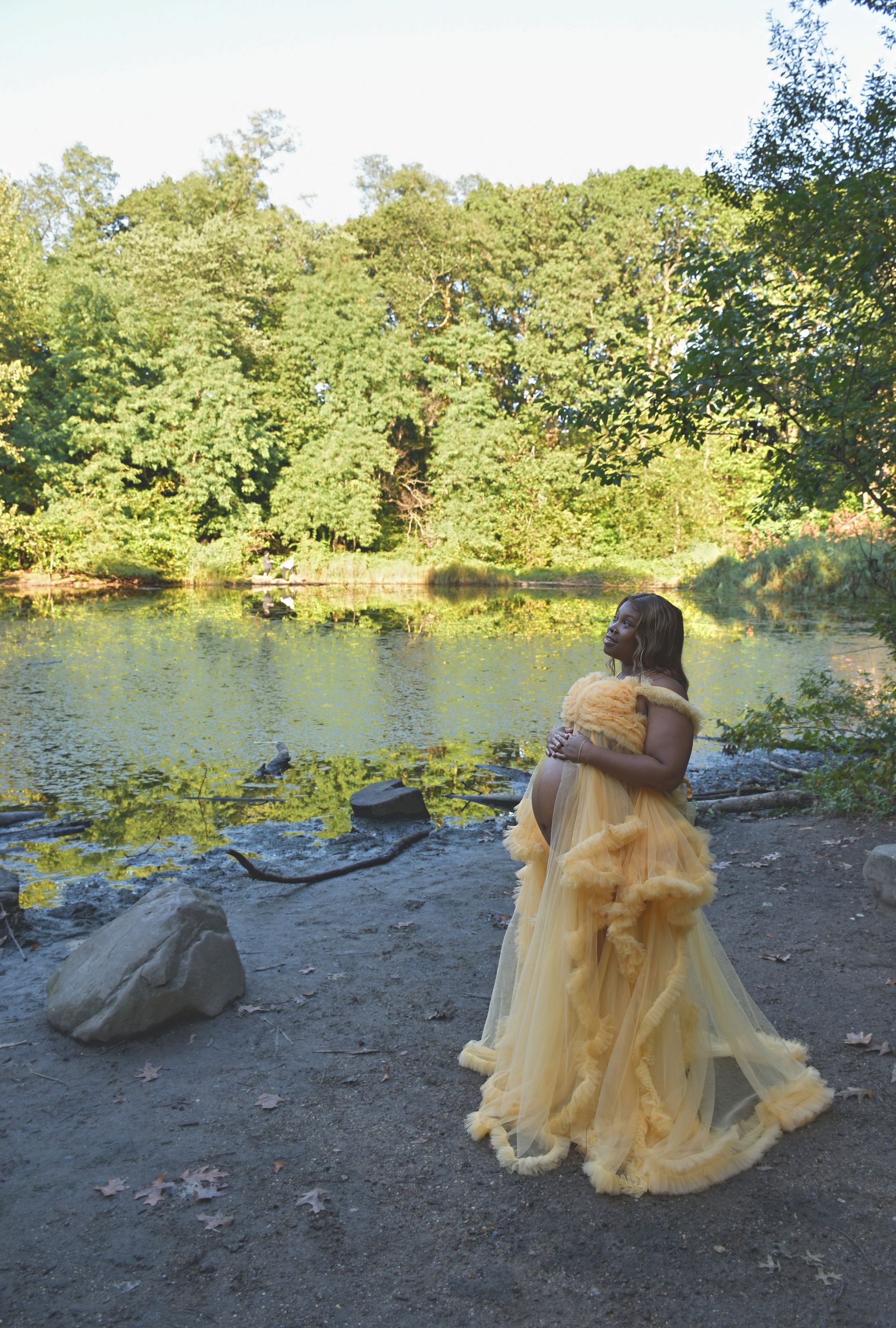 Pregnant person wearing a yellow dress, standing near a lake in a park, looking up.