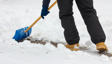 Person shoveling snow from a sidewalk, wearing black pants, tan boots, and blue gloves.