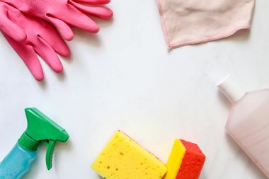 Cleaning supplies arranged on a white surface: pink gloves, spray bottle, sponges, and a cloth.