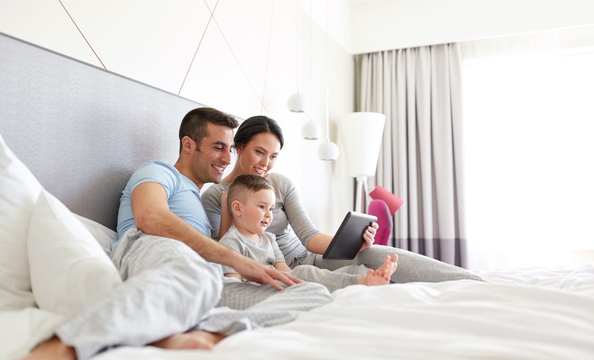 Family sitting on a bed together, using a tablet in a bright modern bedroom.