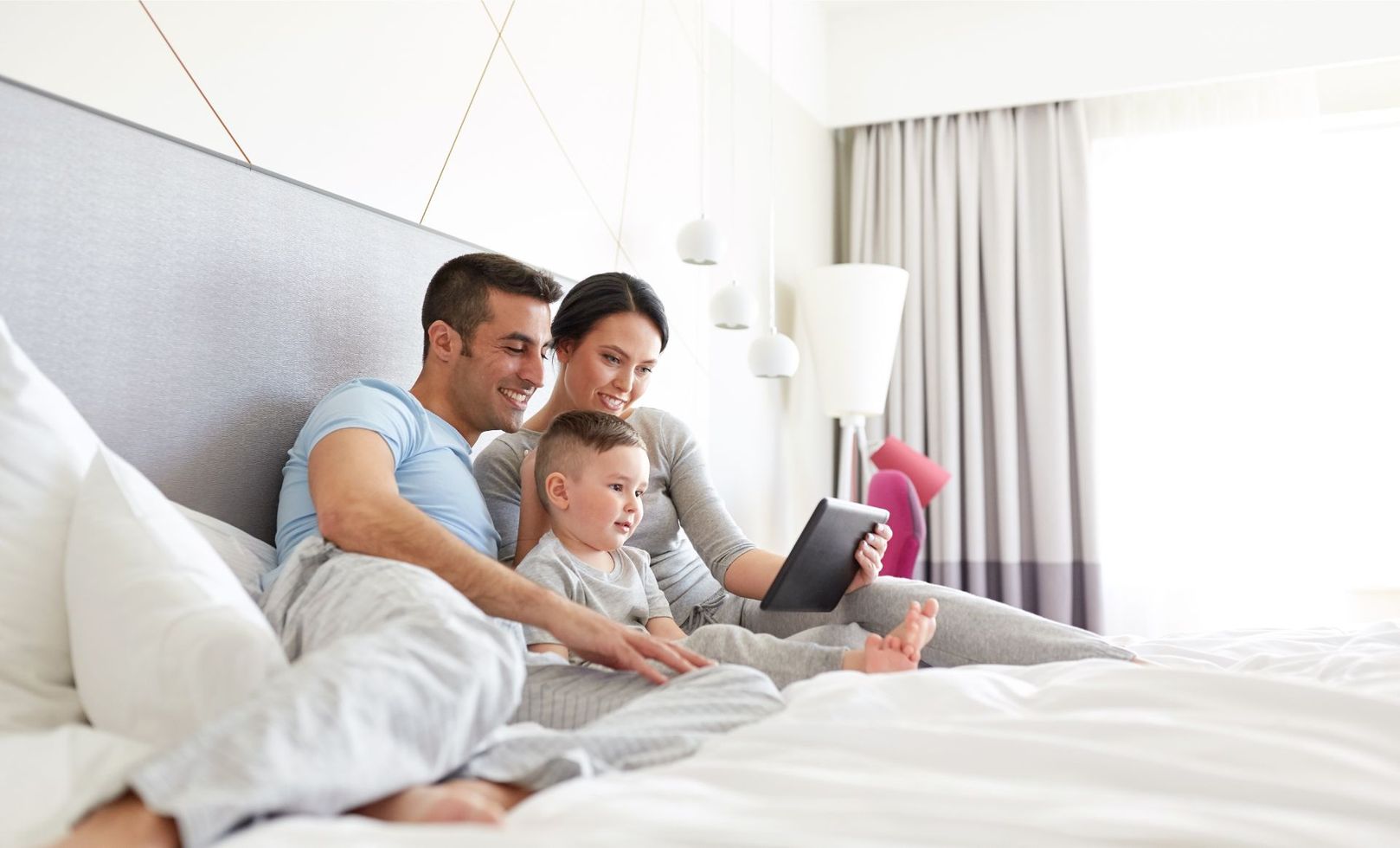 Family sitting on a bed together, using a tablet in a bright modern bedroom.