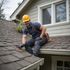 Roofer in hard hat on a roof, inspecting shingles. He smiles, wearing work gloves and a tool belt.