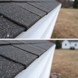 Close-up of a roof's shingles and a white gutter, with a blurred background of a yard and a house.