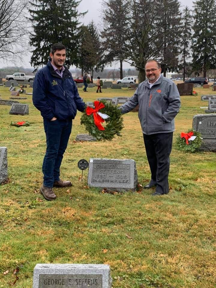 Two men are standing in a cemetery holding a wreath.