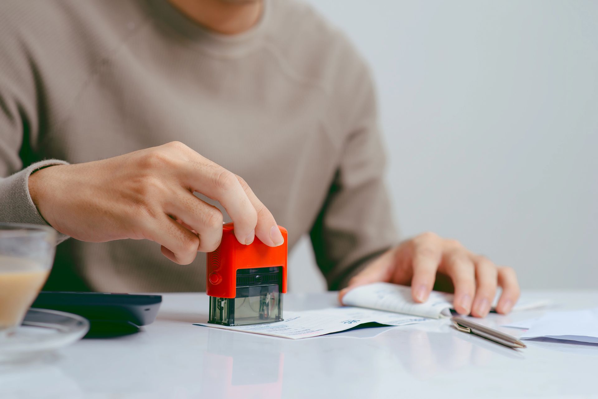 Una persona estampa un documento con un sello rojo y negro en un escritorio. Lleva un suéter color canela y hay un bolígrafo y un café cerca.