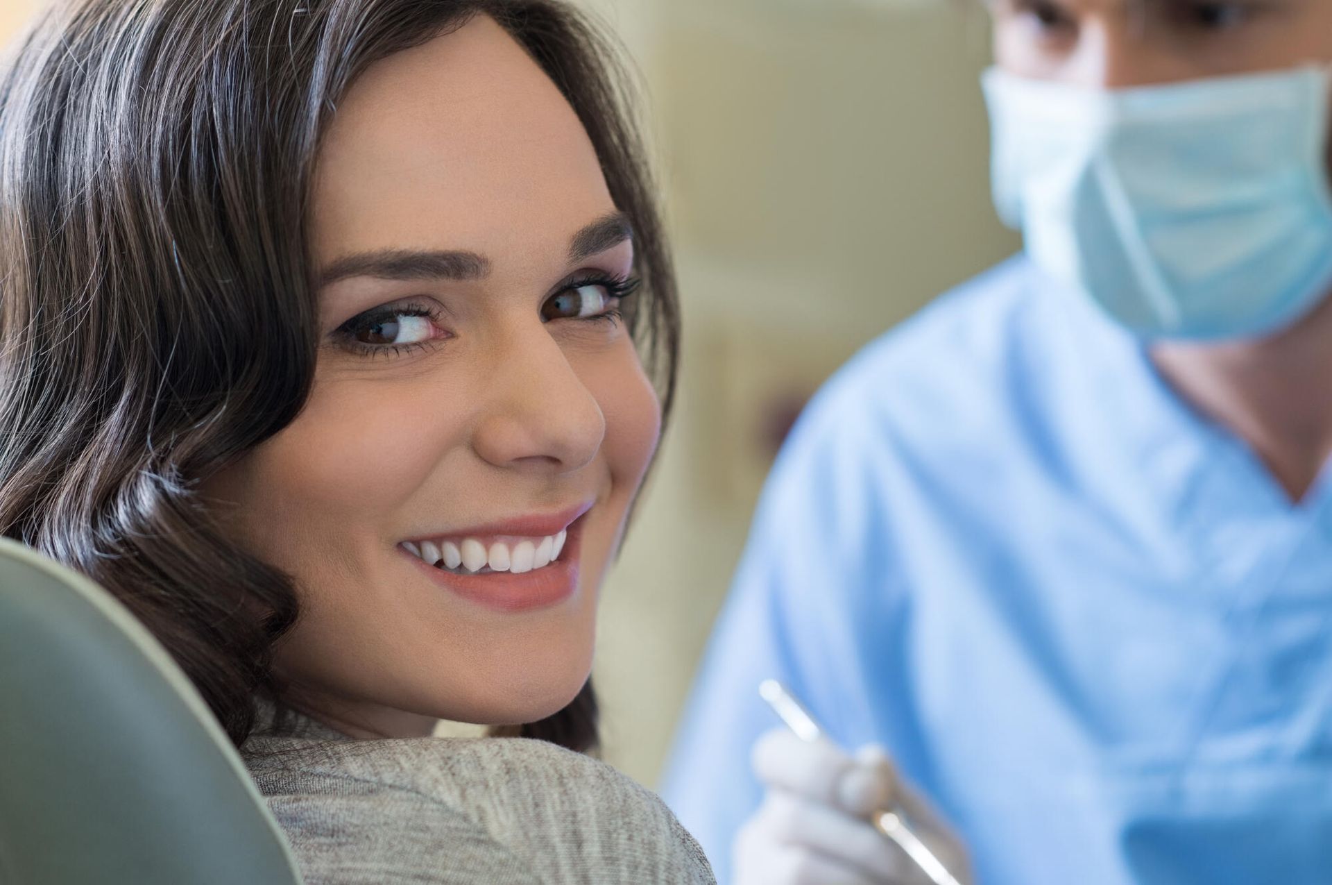 A woman is smiling while sitting in a dental chair while a dentist examines her teeth.
