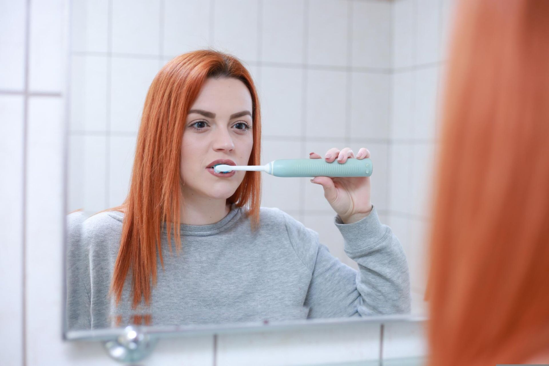 Woman brushing teeth in a bathroom mirror, holding a toothbrush near her mouth.