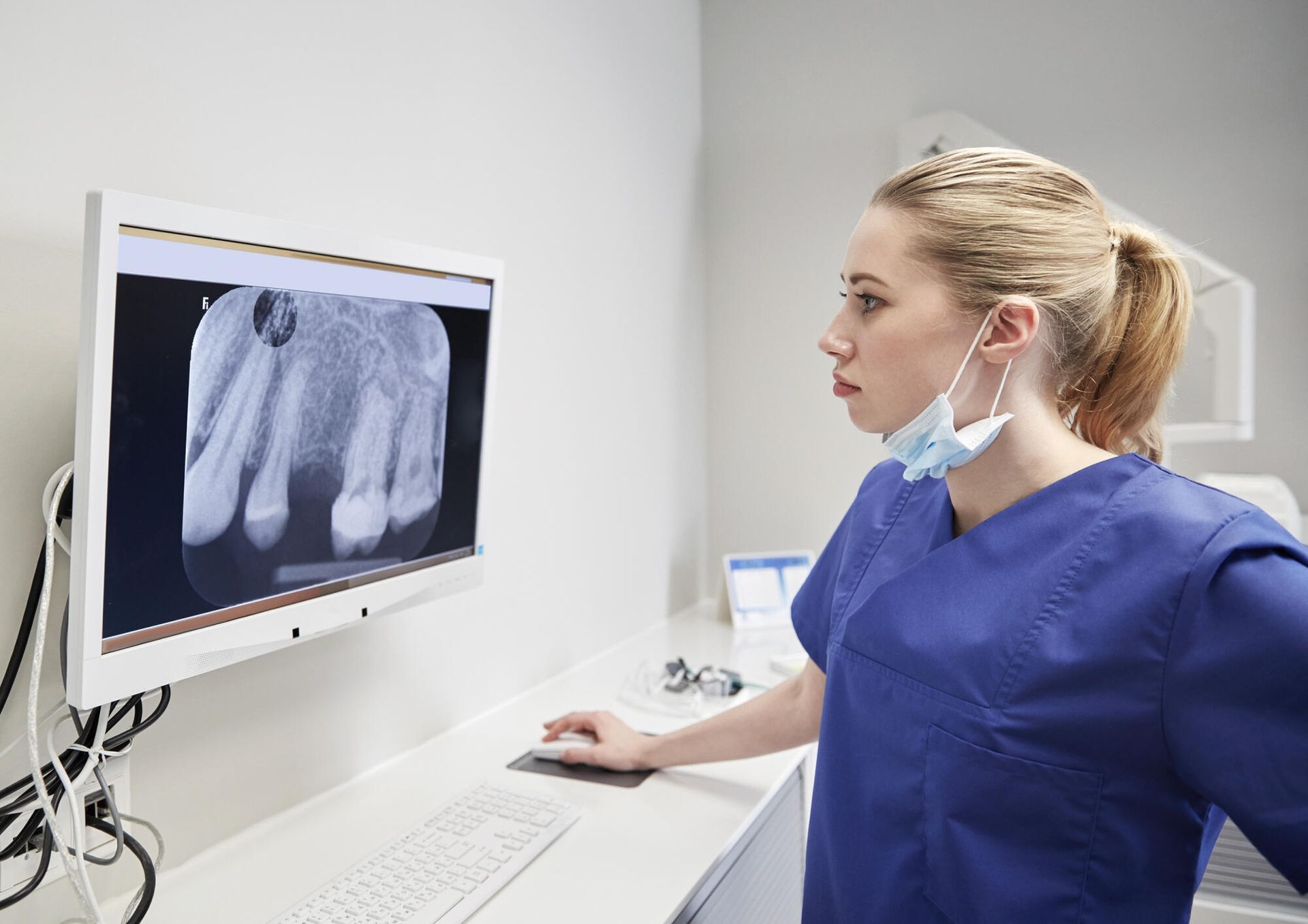Dentist in blue scrubs examines a tooth X-ray on a monitor in a dental office.