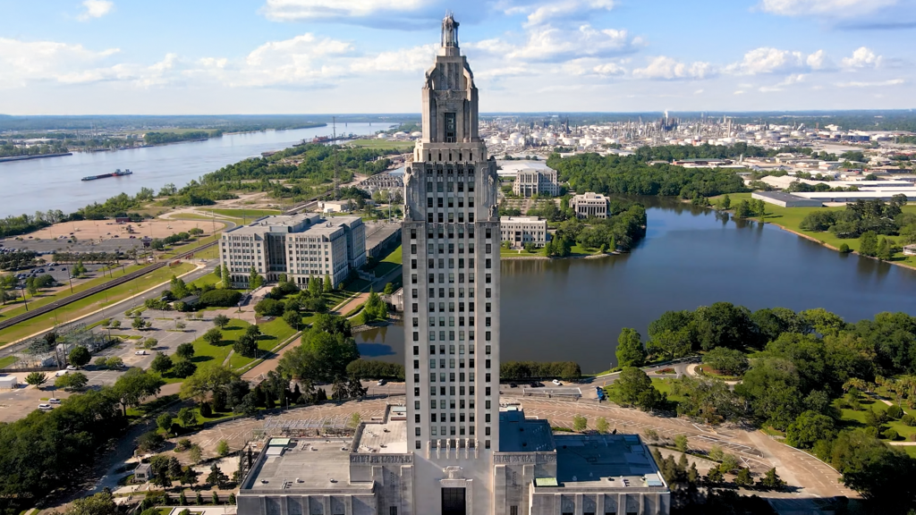Louisiana State Capitol building against the backdrop of a city and river, under a blue sky.