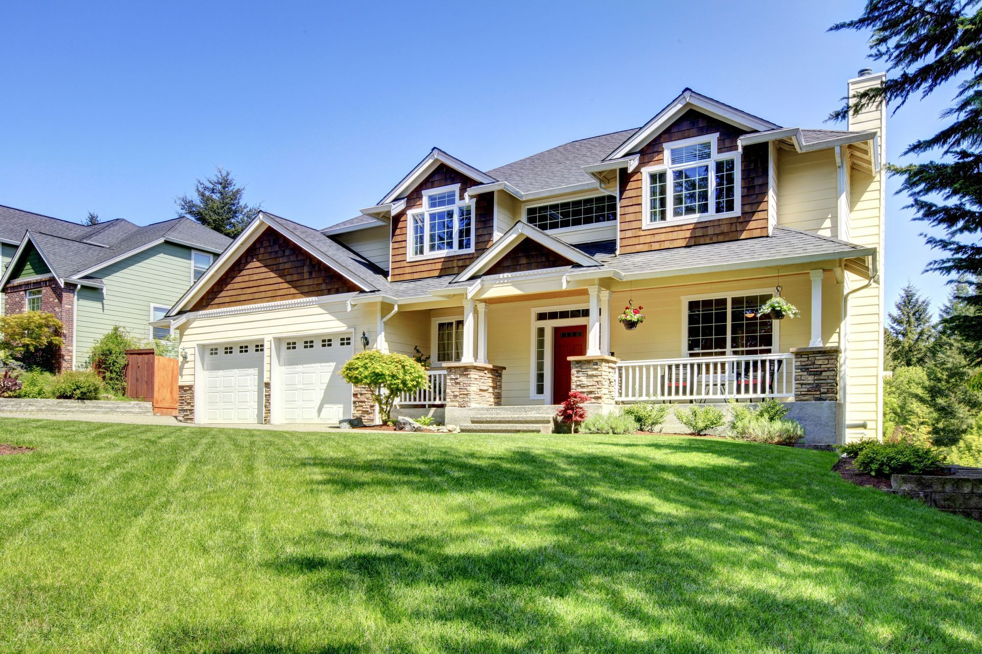 Two-story yellow house with brown accents, white porch, and garage on a green lawn under a blue sky.