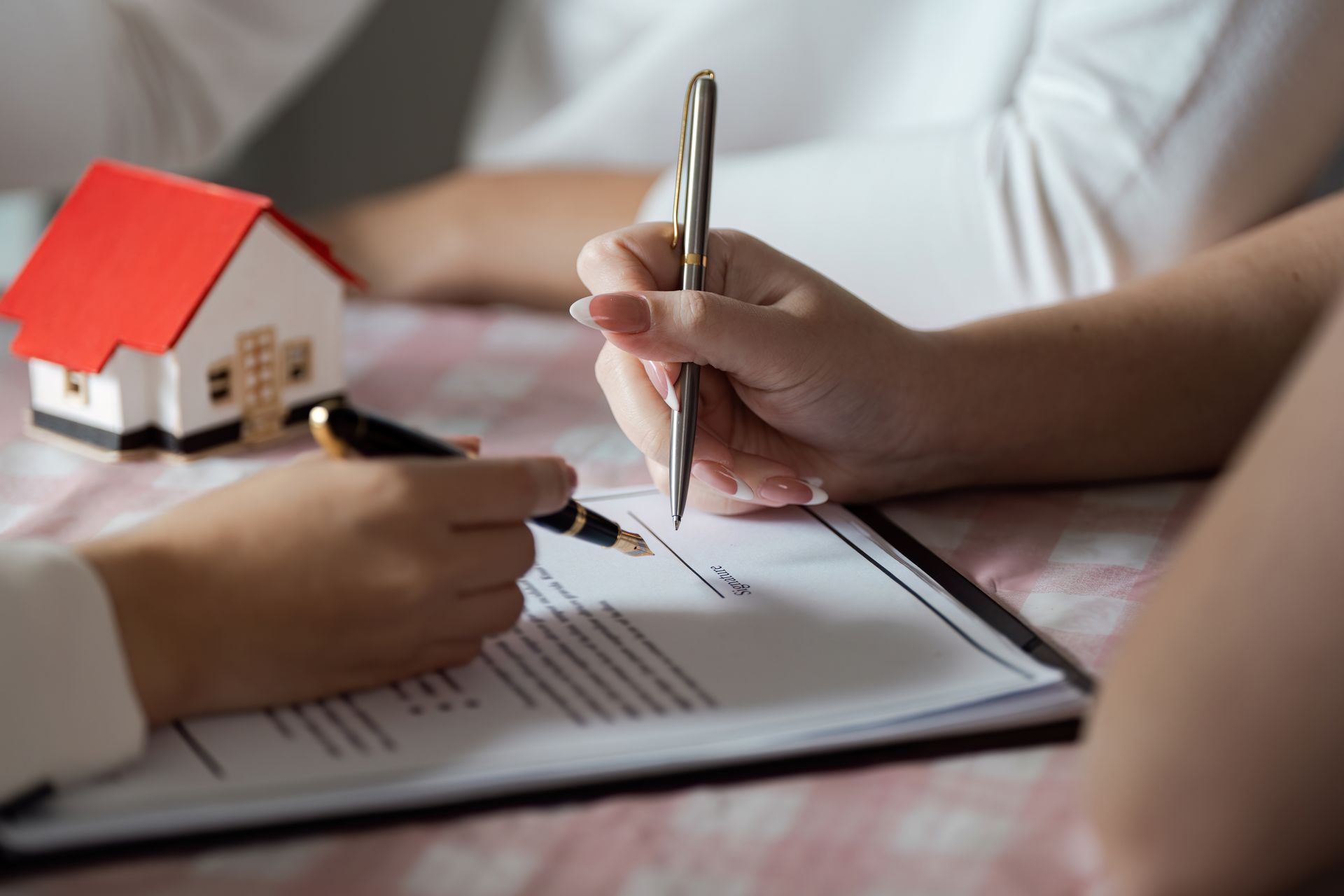Person signs a real estate document beside a small model house on a desk. Person signs a real estate document beside a small model house on a desk.