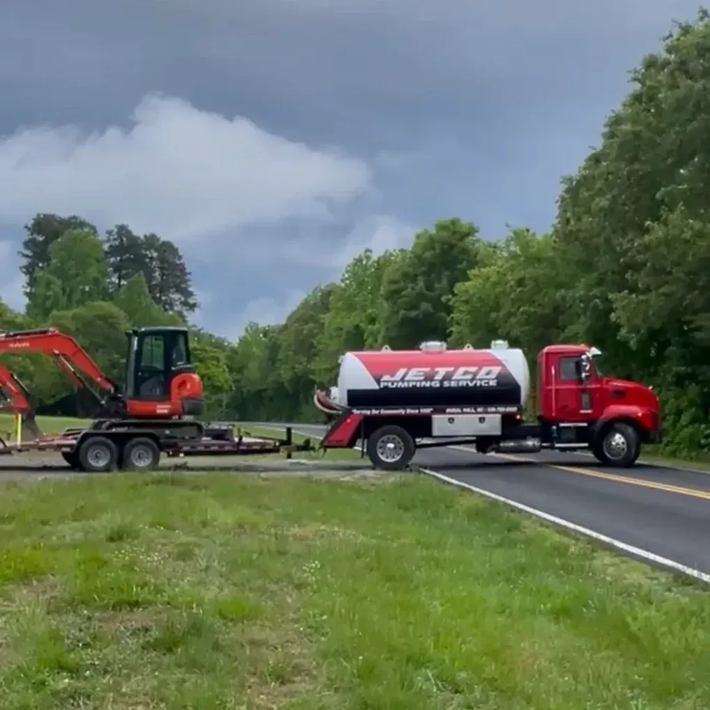 Red tanker truck towing an orange excavator on a rural road under cloudy skies