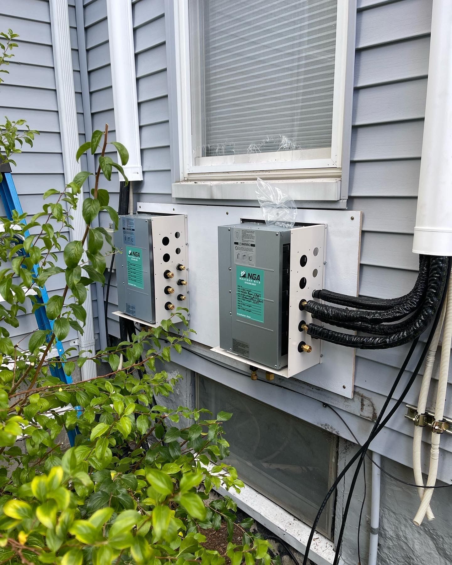 Two silver utility boxes mounted on a white board outside a house. Wires and pipes extend from the boxes.