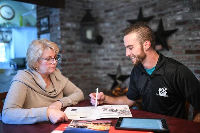 A man shows a woman a document. They sit at a table, likely discussing home improvement, with a tablet nearby.