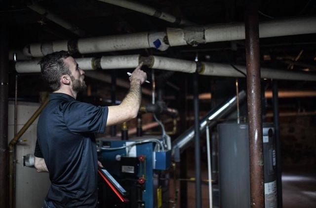Man inspecting pipes in a basement, pointing with a flashlight.