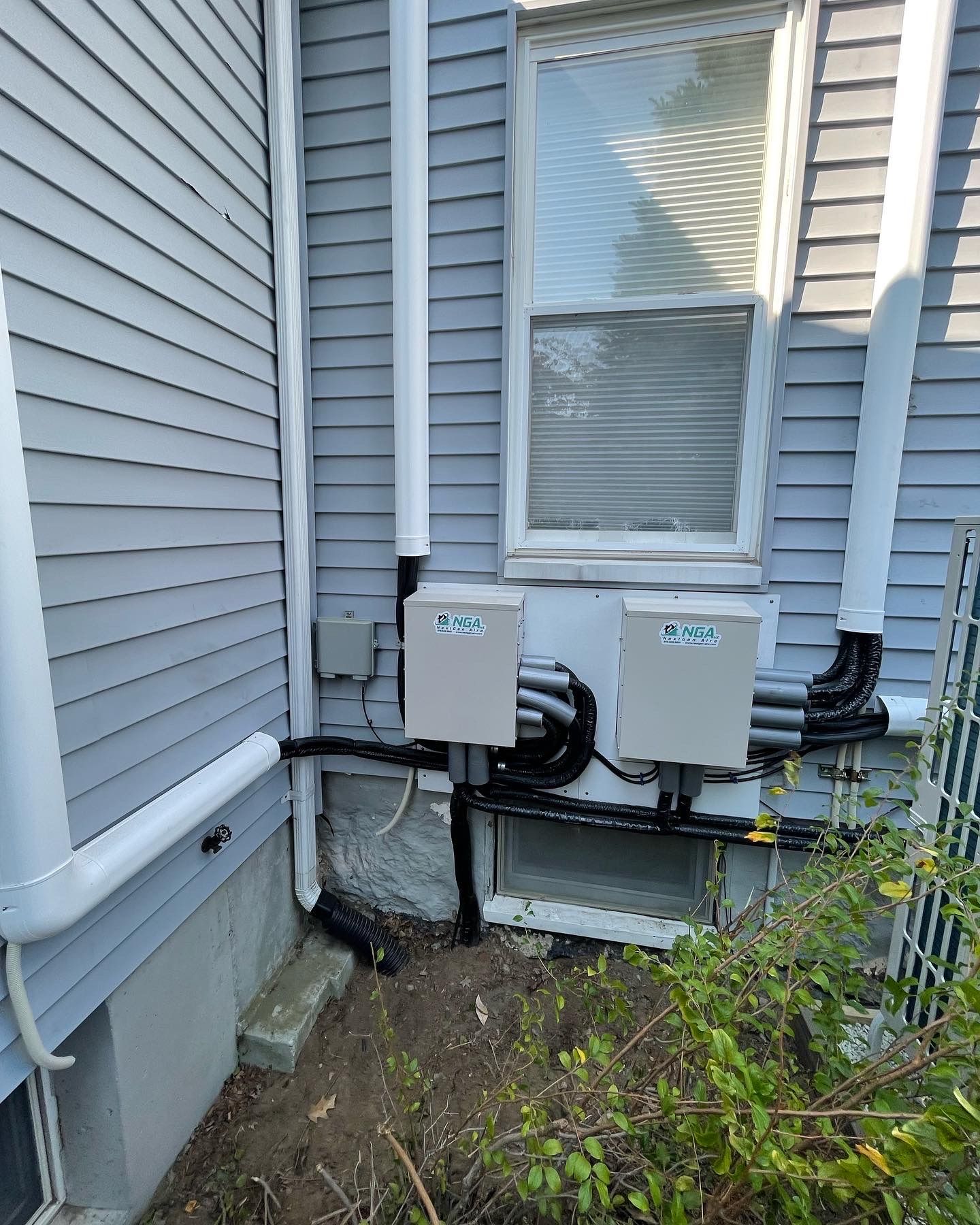 Grey siding with white pipes and two gray boxes mounted near a window. Black conduits connect the boxes.