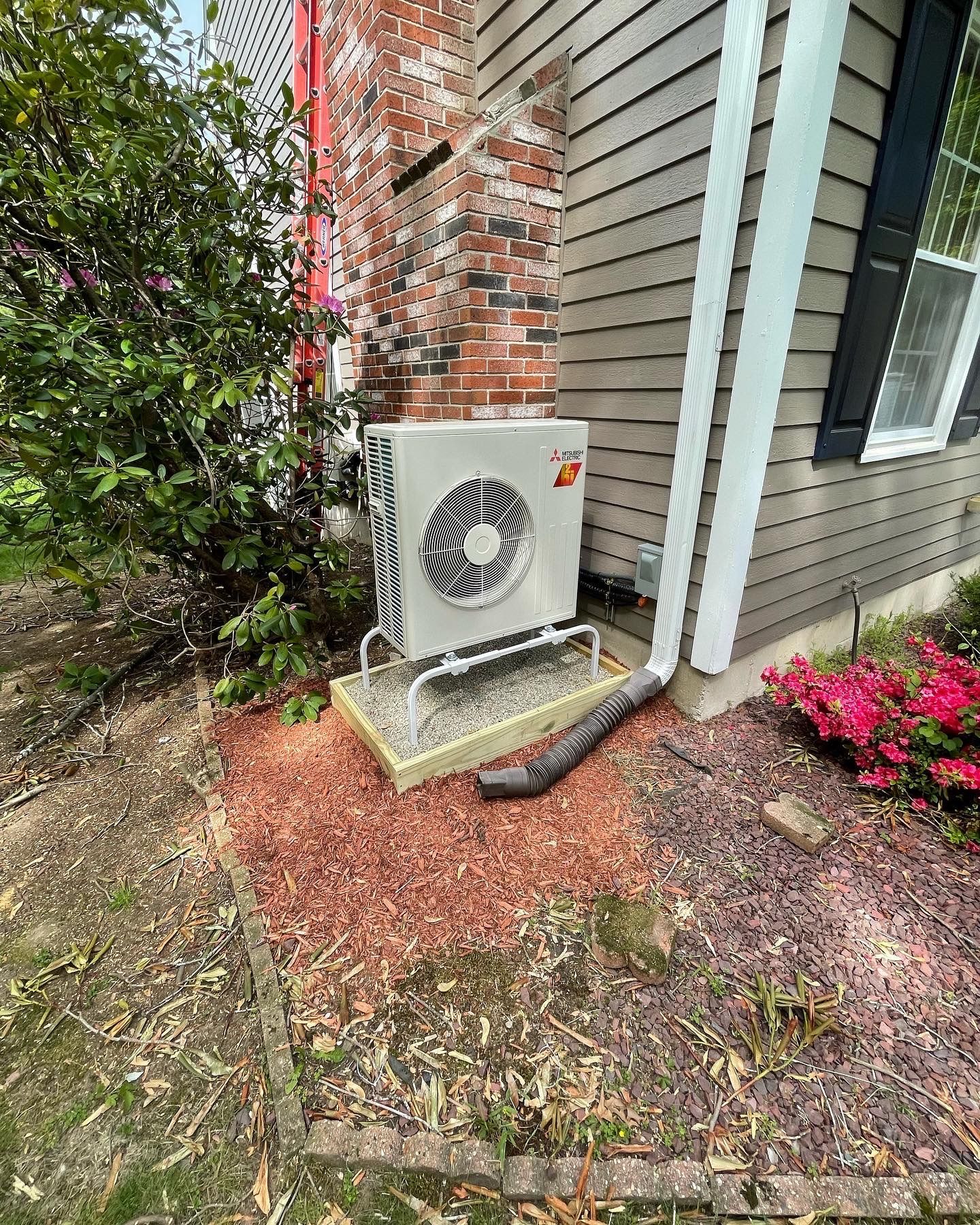 Outdoor HVAC unit next to brick chimney and house with brown siding, gravel base, and red mulch.