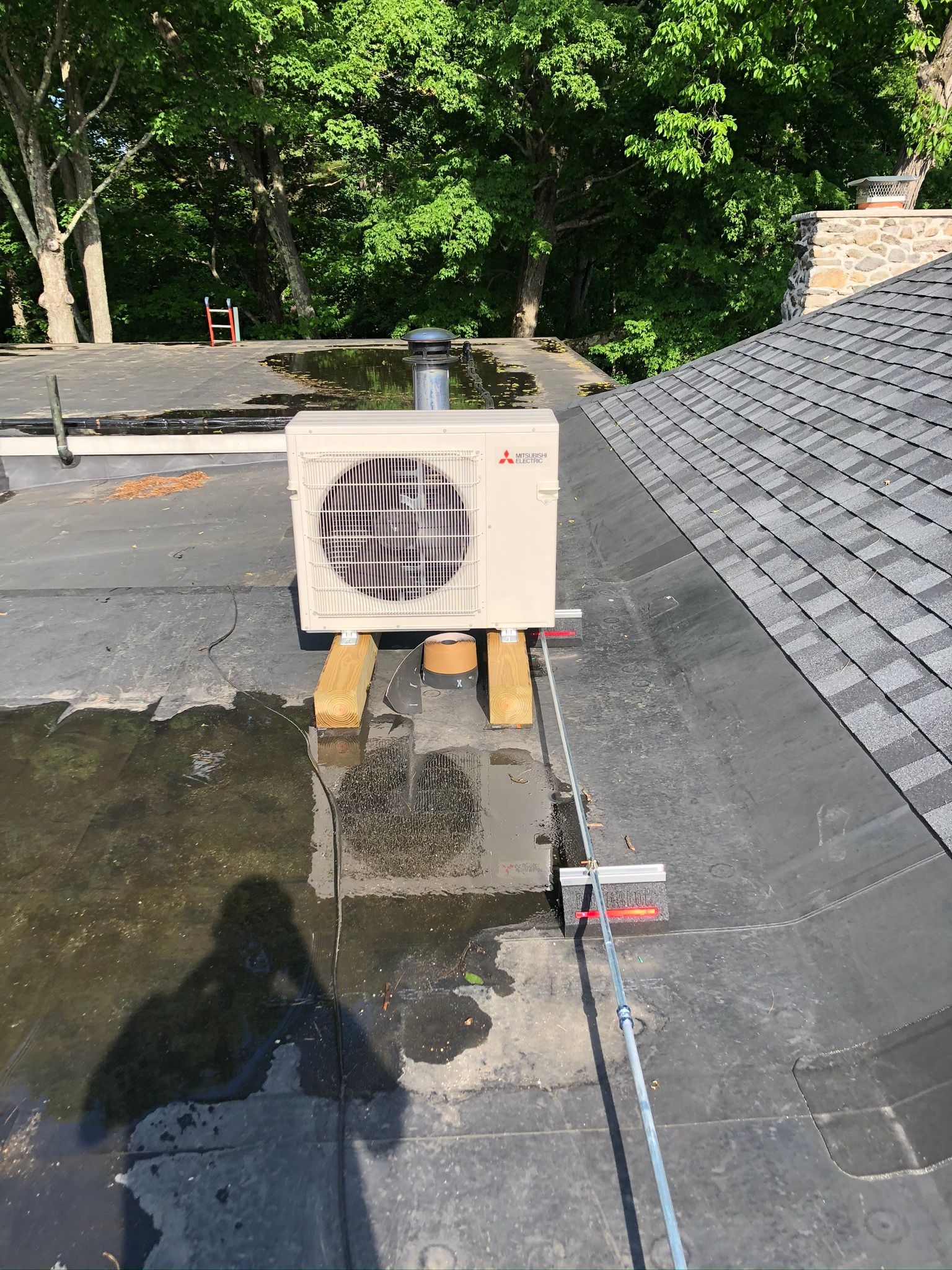 Mini-split AC unit on wooden supports, rooftop. Water pools around it; trees in background.