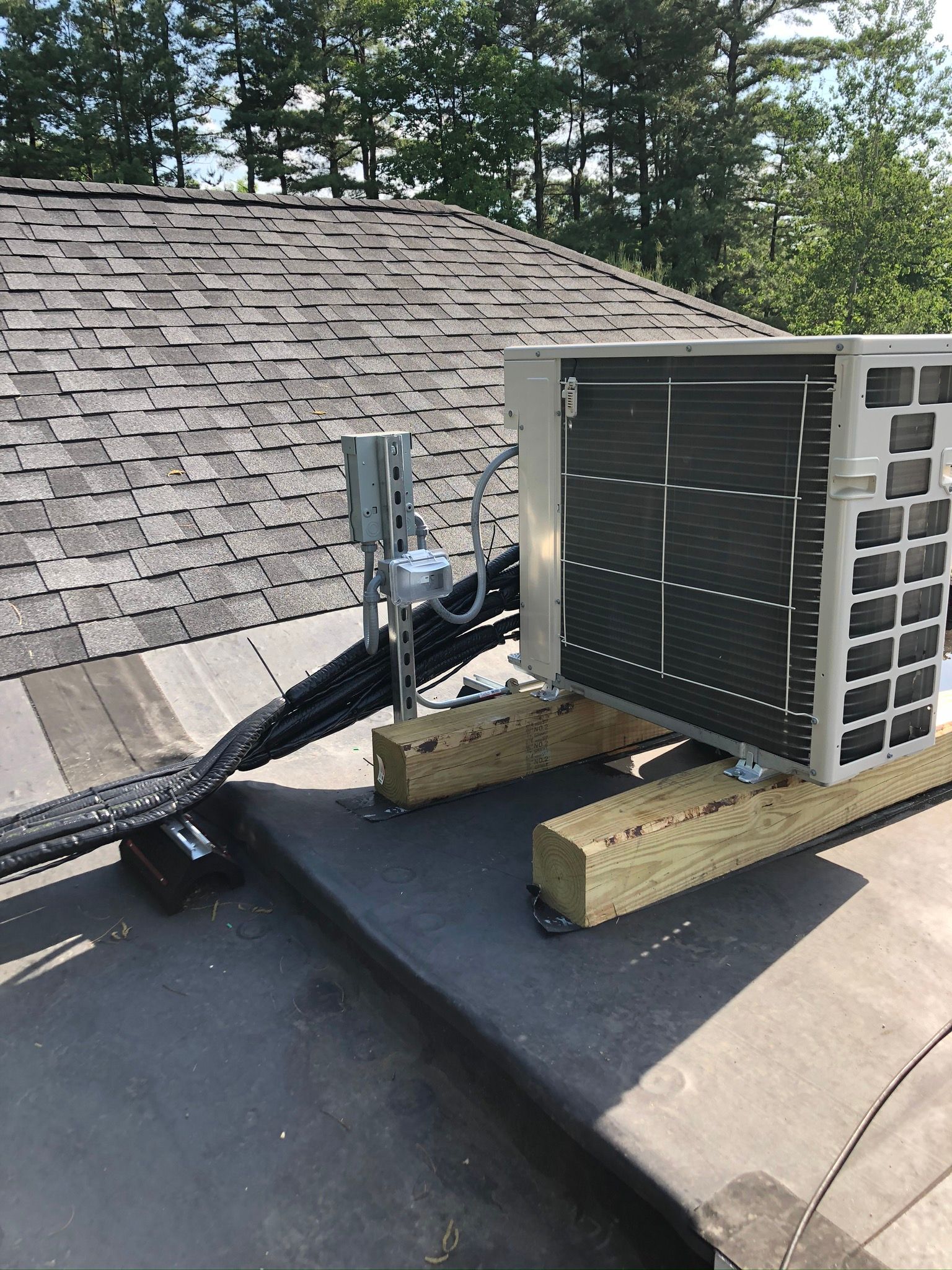 Air conditioning unit on wooden beams on a flat roof with black conduit and grey shingles.