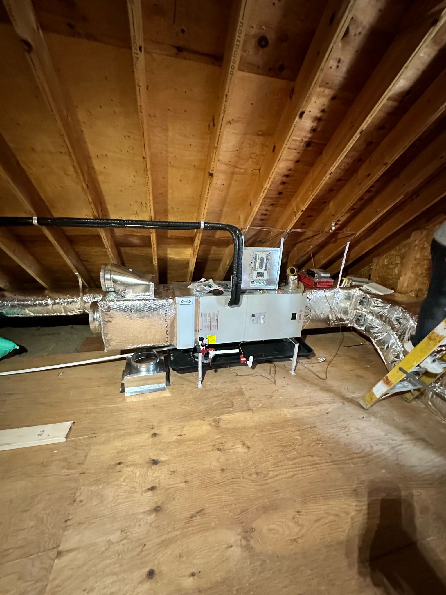 HVAC unit in an attic with wooden beams and plywood flooring. Insulation lines the walls.