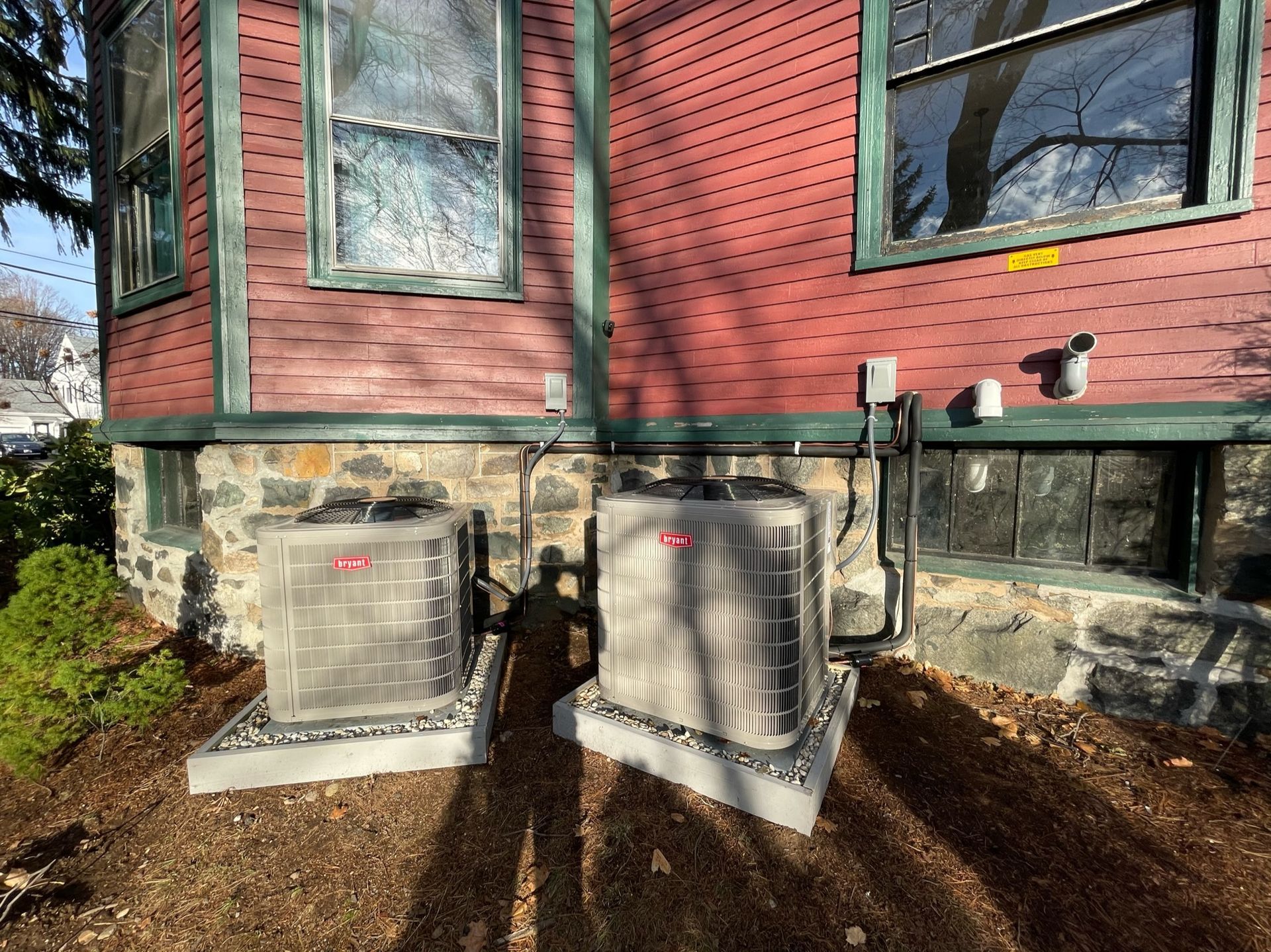 Two air conditioning units sit on gravel pads next to a red house with windows.