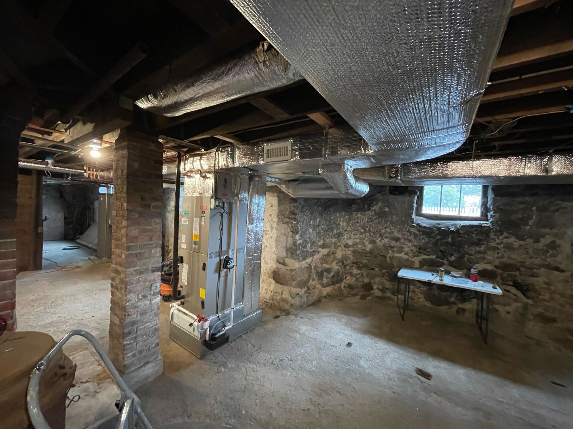 Basement with exposed brick columns, ductwork, and HVAC unit. Raw stone walls and a small window are visible.