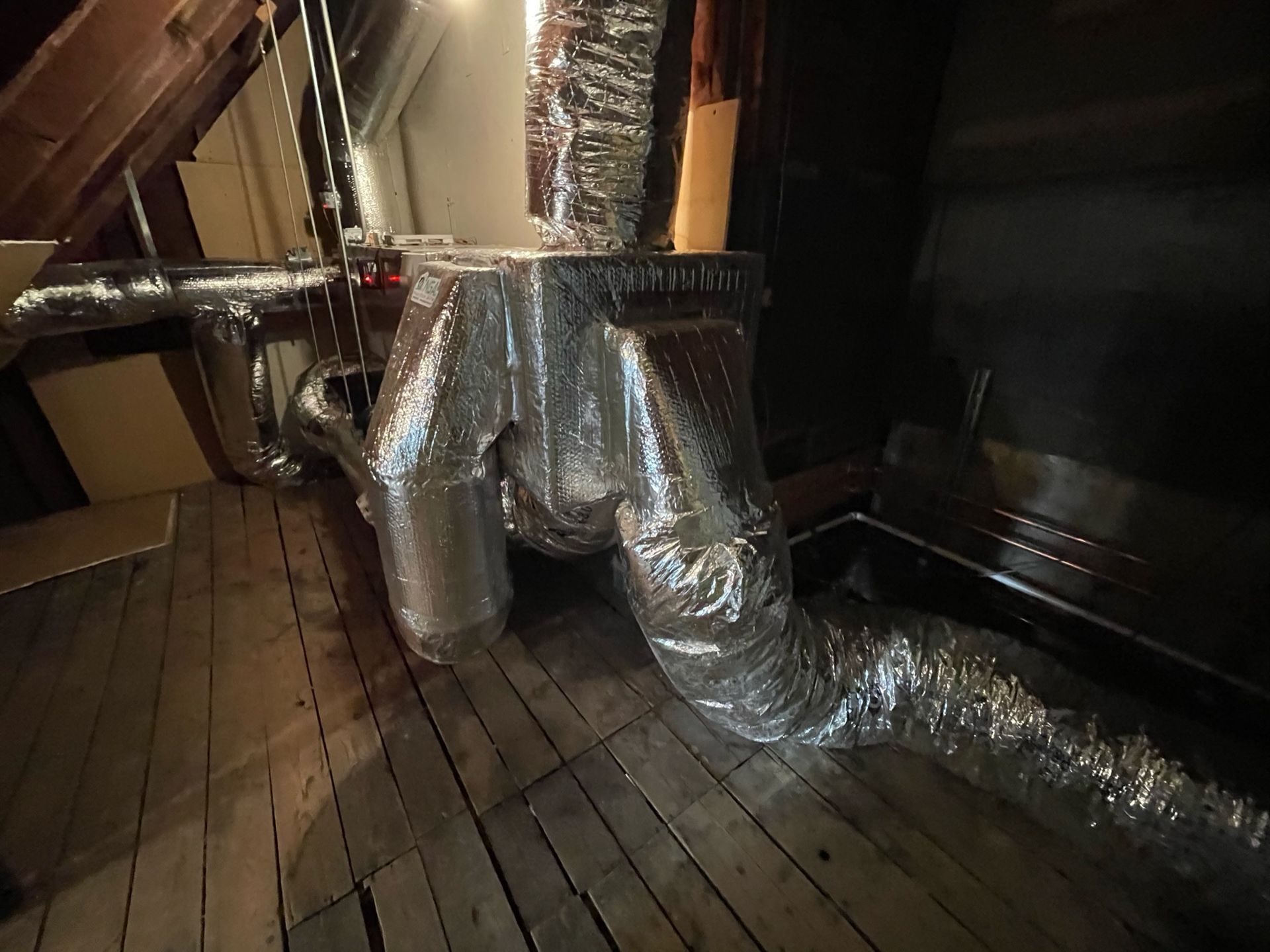 HVAC ductwork in an attic, with metallic, flexible ducting. Wooden floor and dark interior.