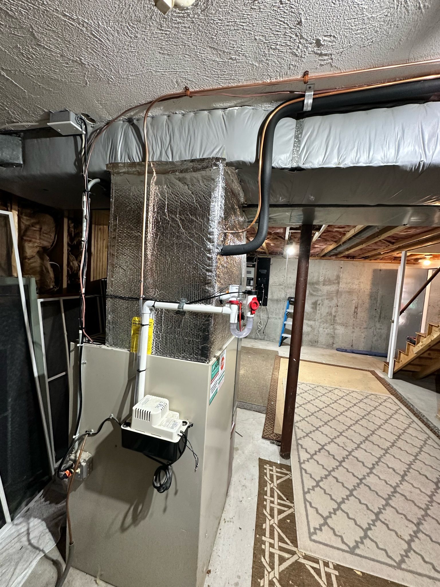 Basement utility area with furnace, ducts, and pipes against concrete walls and a patterned rug.