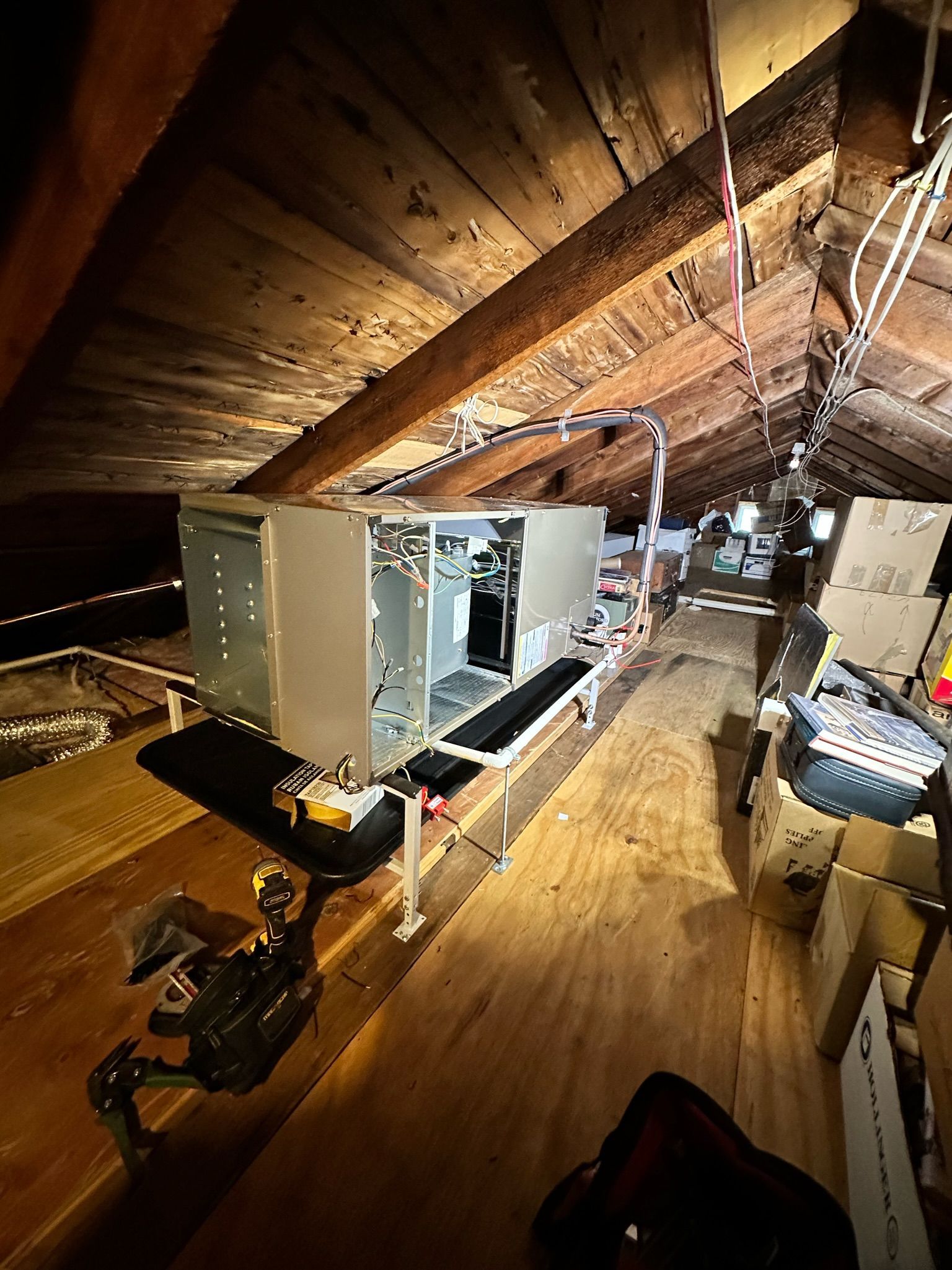 HVAC unit in an attic with wooden beams and a narrow wooden walkway. Wiring and various items are scattered around.