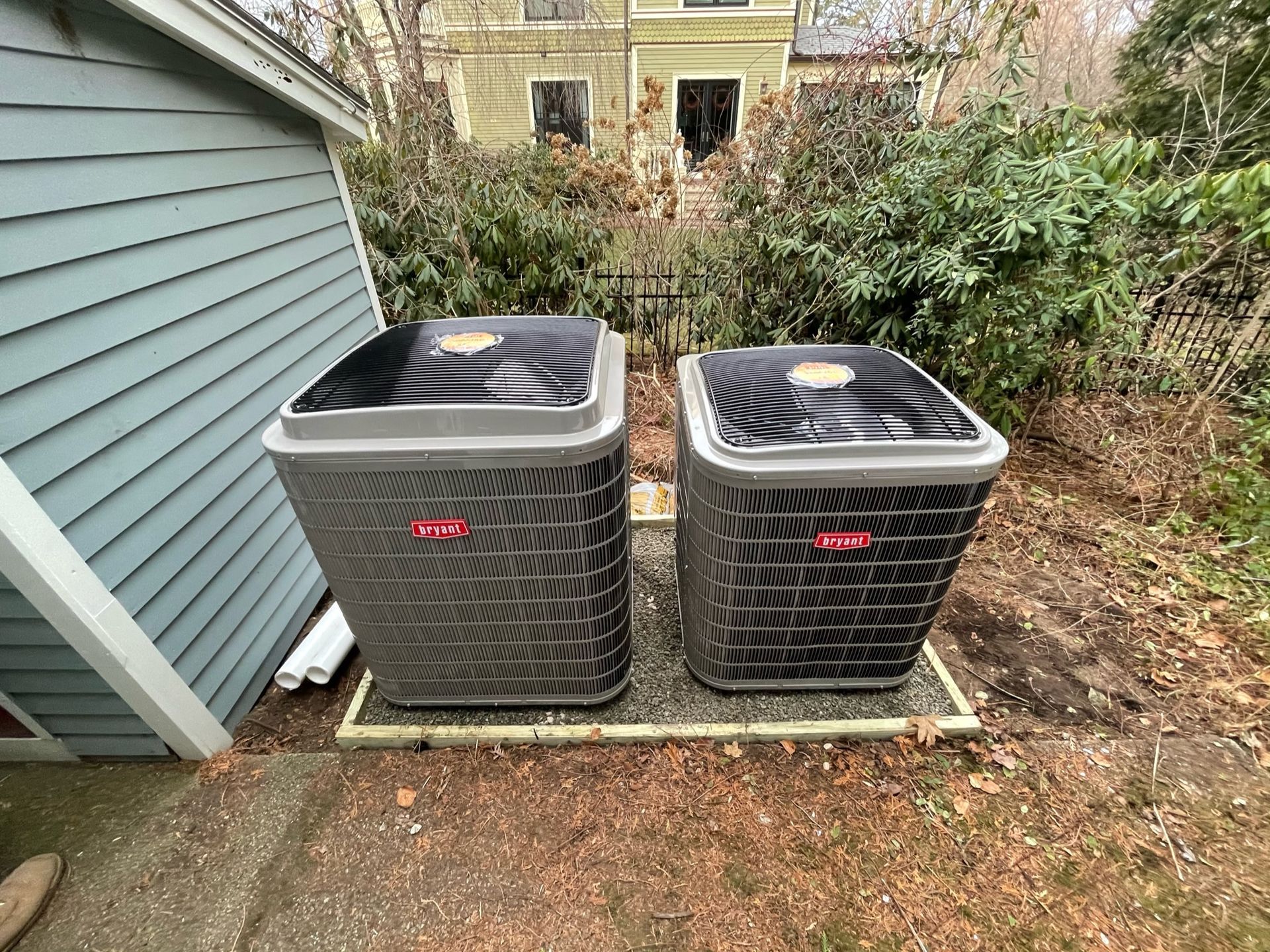 Two dark-colored air conditioning units on a gravel base next to a blue building and greenery.