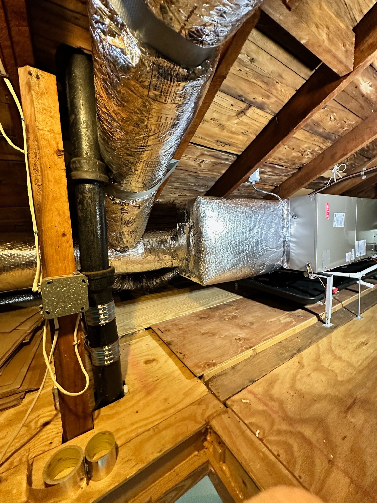 Attic space with ductwork and a dark pipe. Wooden beams and plywood flooring visible.
