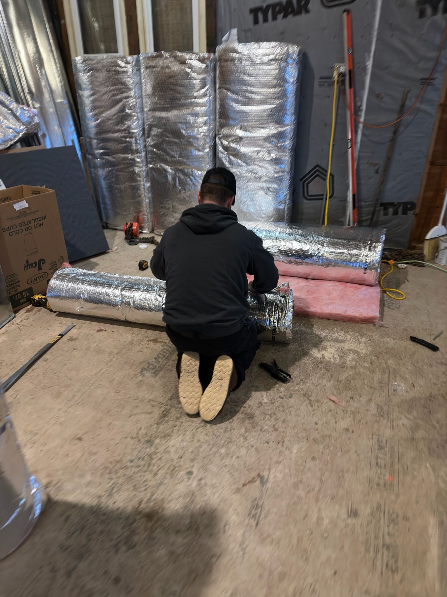 Person installing insulated ductwork in a basement, surrounded by materials and tools.