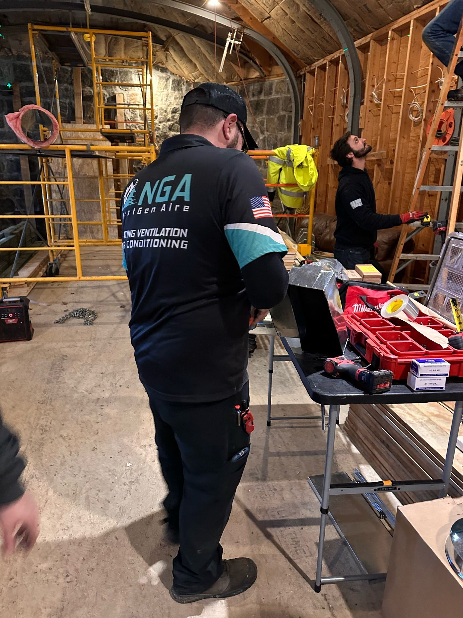 Construction workers in a room with tools; one wears a black shirt with 