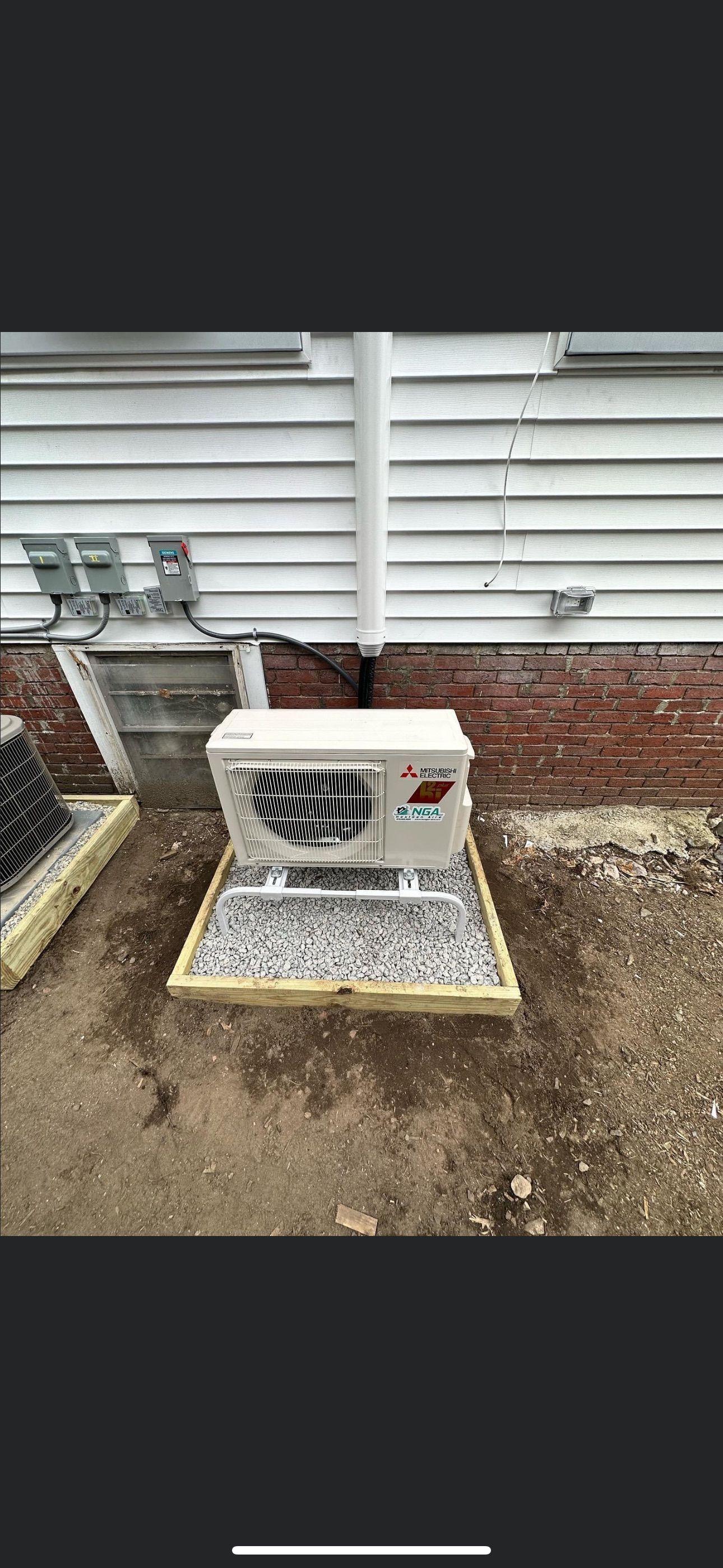 An outdoor air conditioning unit sits on a gravel base next to a white clapboard wall and brick foundation.