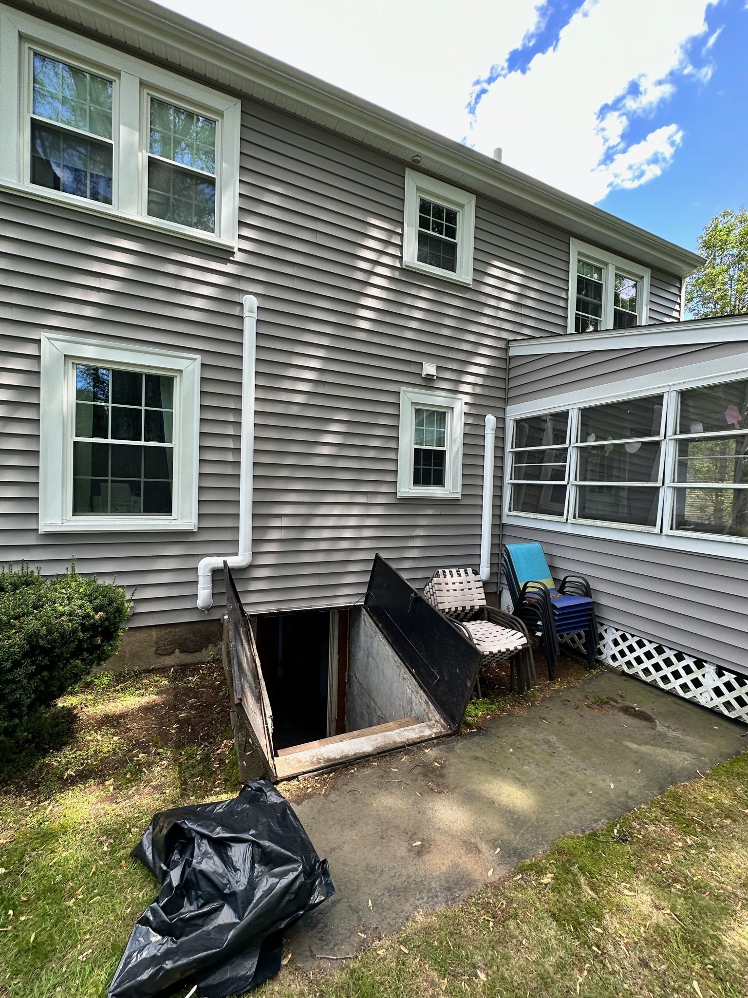 Exterior of a gray house with a basement entrance and a screened porch. A black trash bag sits in the foreground.