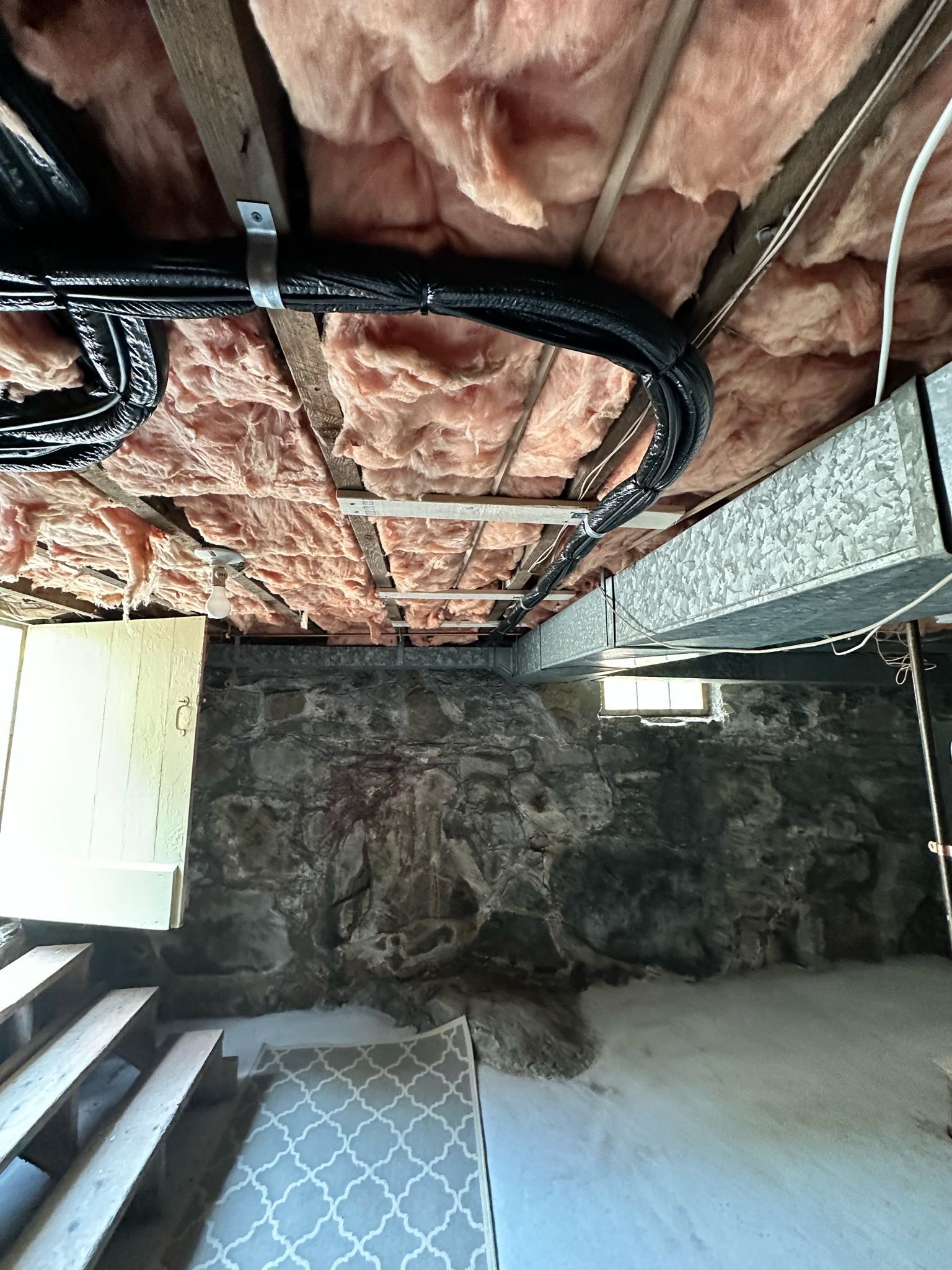 Basement interior with exposed stone wall, wood stairs, ceiling insulation, and electrical wiring.