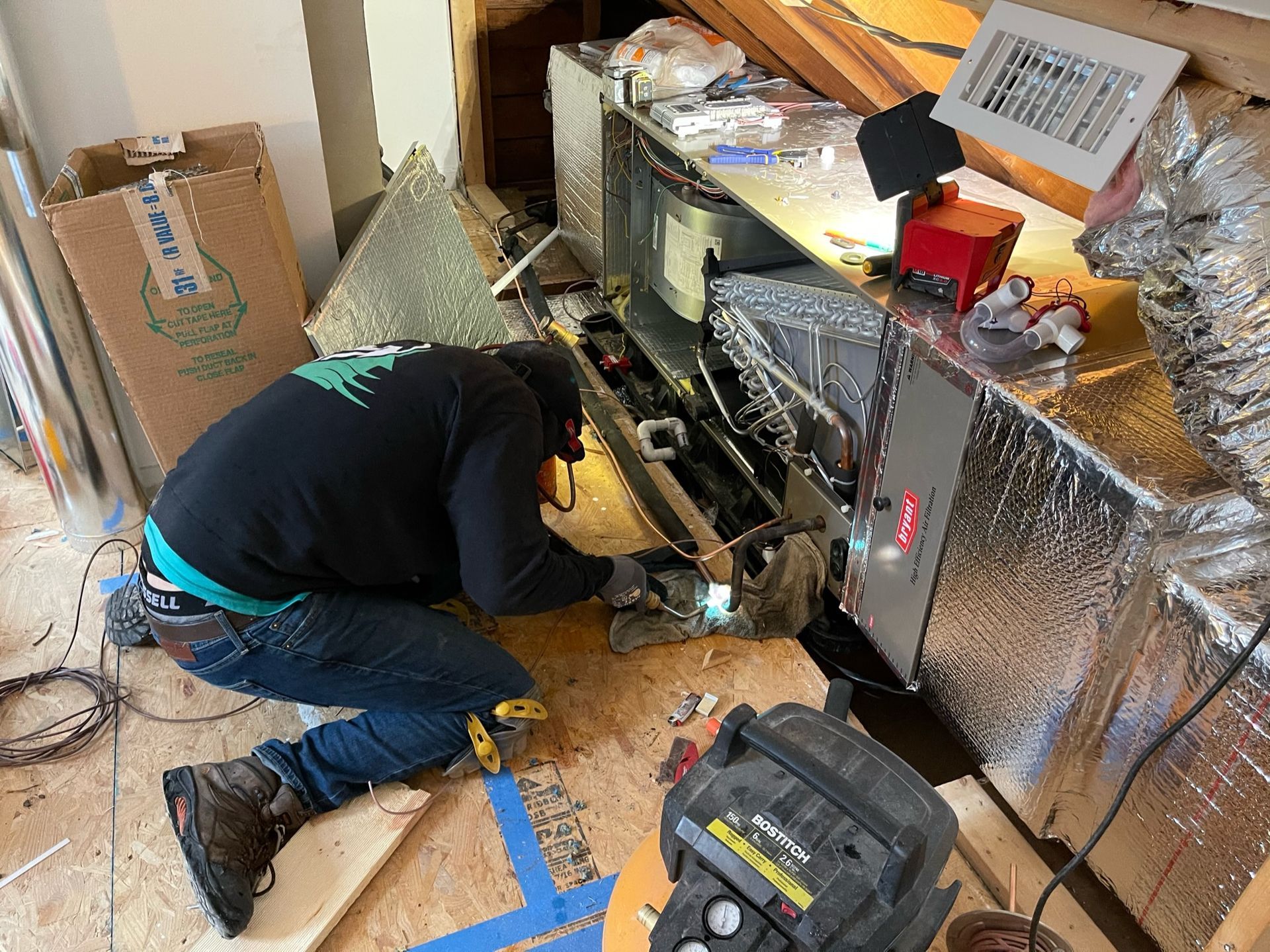 Person works on HVAC unit in an attic, using tools. The unit is silver and insulated, surrounded by building materials.