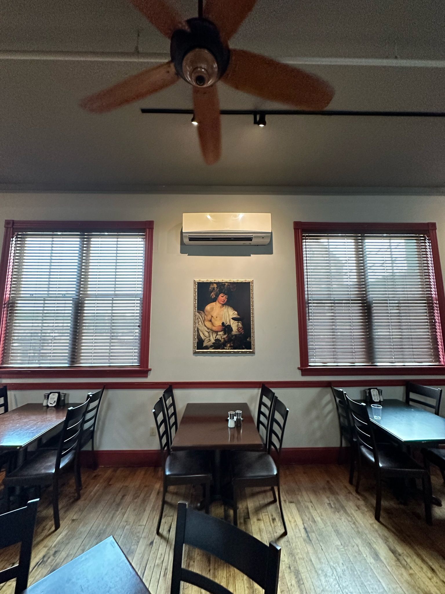 Restaurant interior with tables, chairs, windows with blinds, ceiling fan, and art on the wall.