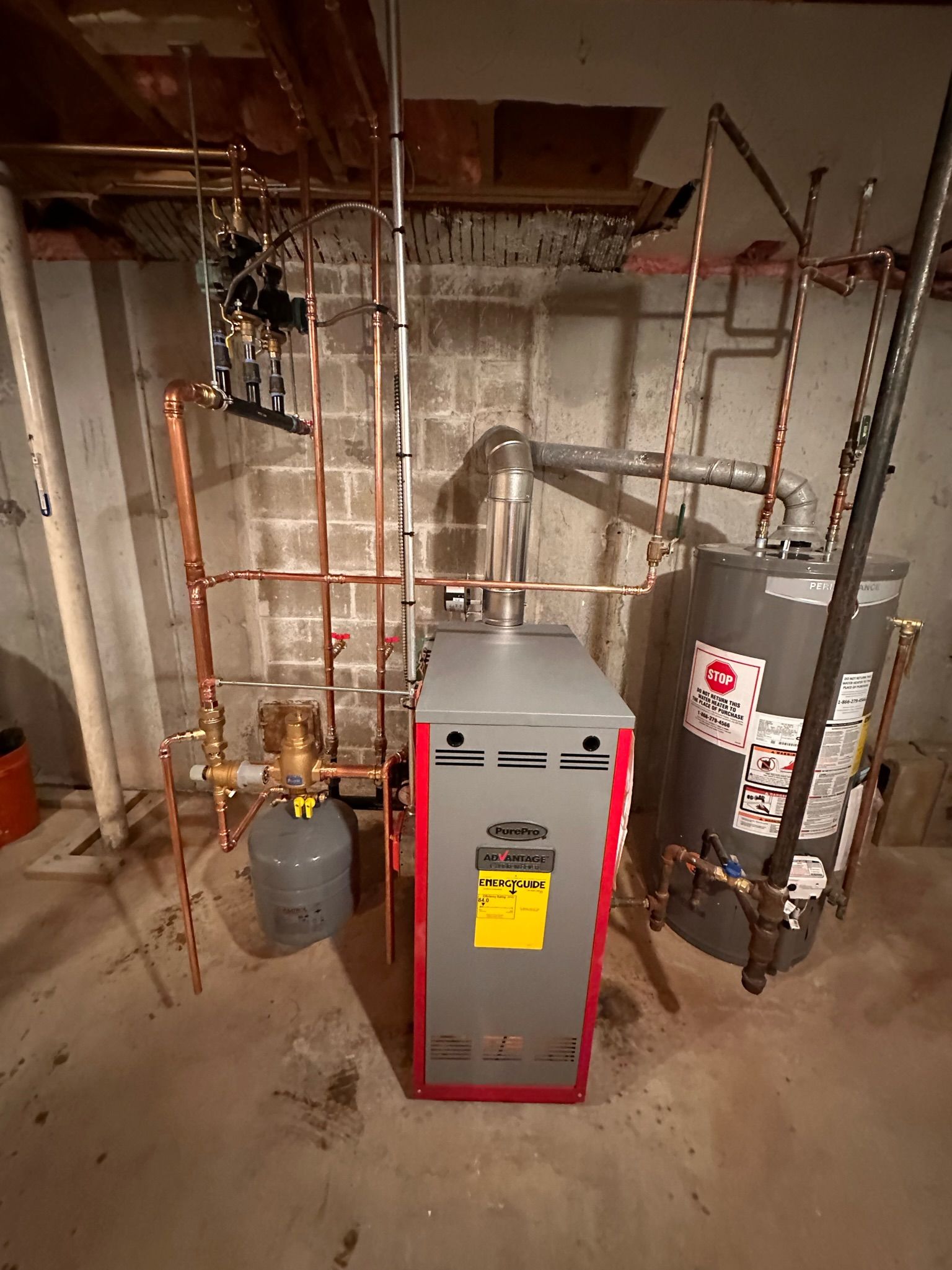 Furnace, water heater, and piping in a basement. Copper and steel pipes are visible with gray and red equipment.