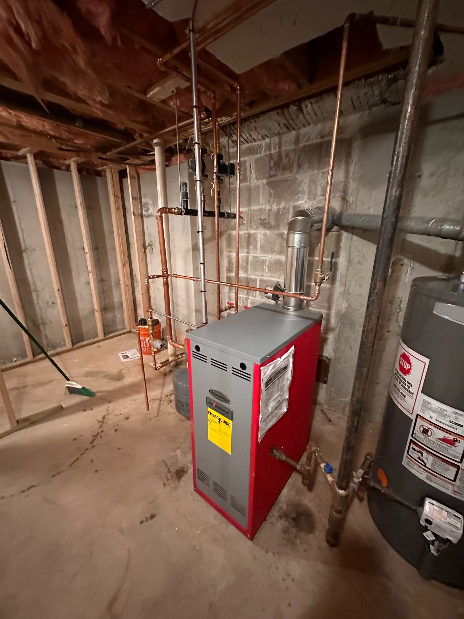 A utility room with a red and gray boiler, copper pipes, and a water heater. Concrete and wooden walls.