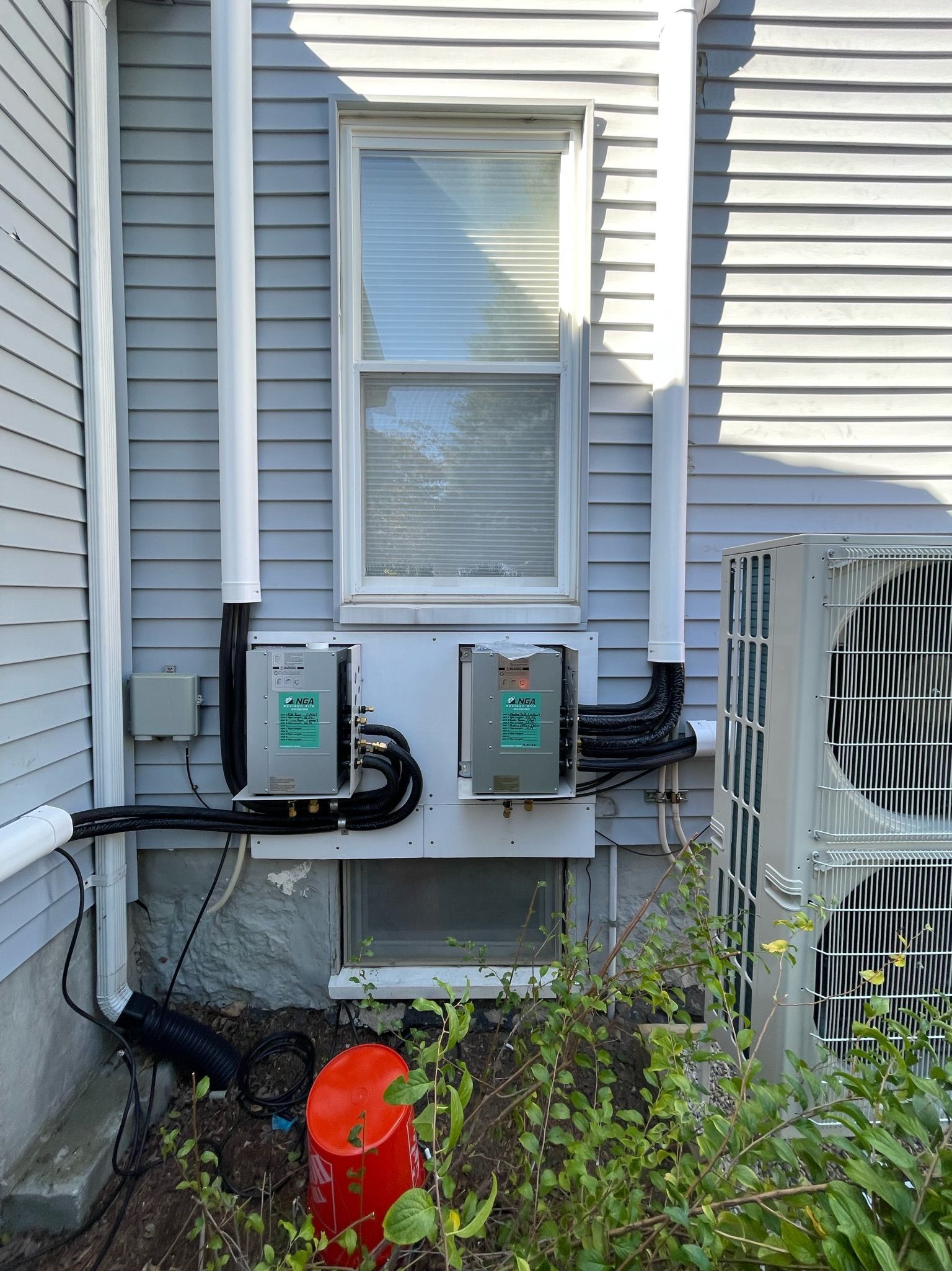 Exterior view of a house with two air conditioning units and a window. White pipes run up the wall.
