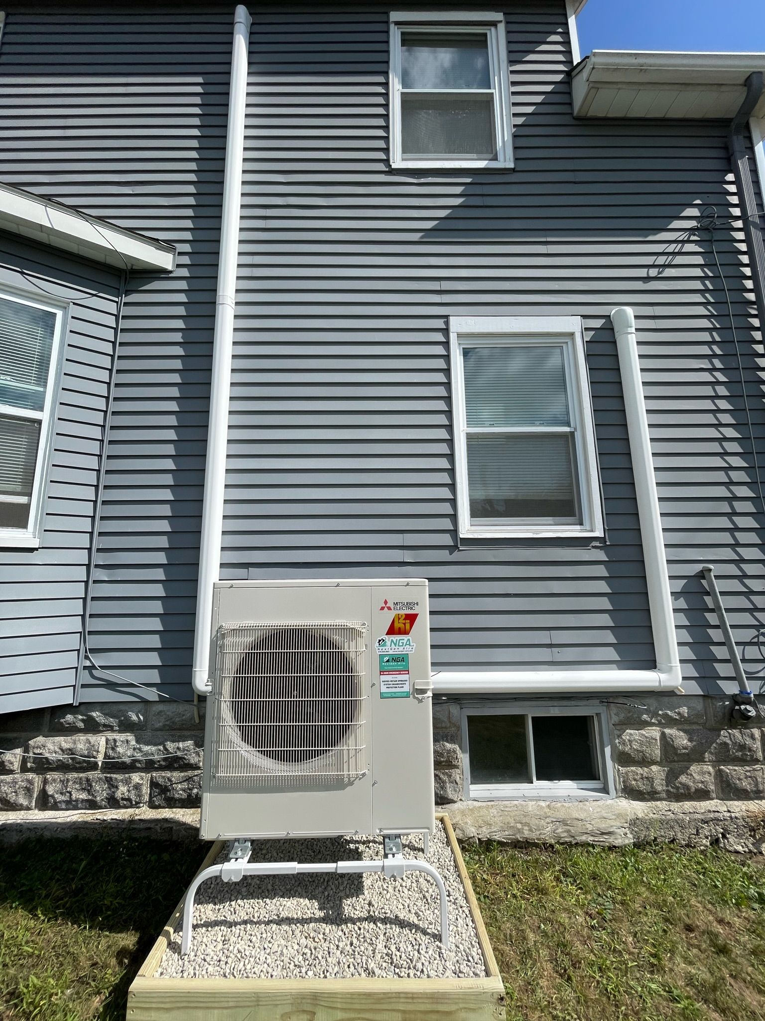 Gray siding on a house with an air conditioning unit on a gravel base. White pipes run up the wall.