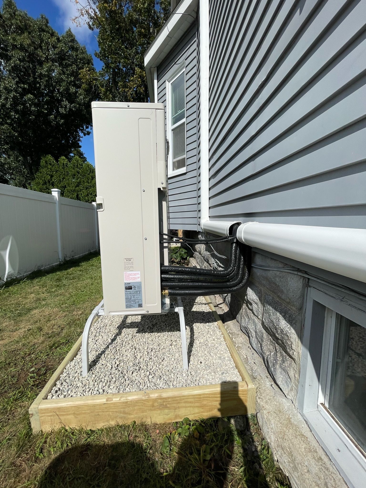 Exterior view of a house with gray siding, an AC unit on gravel, and a white fence in the background.