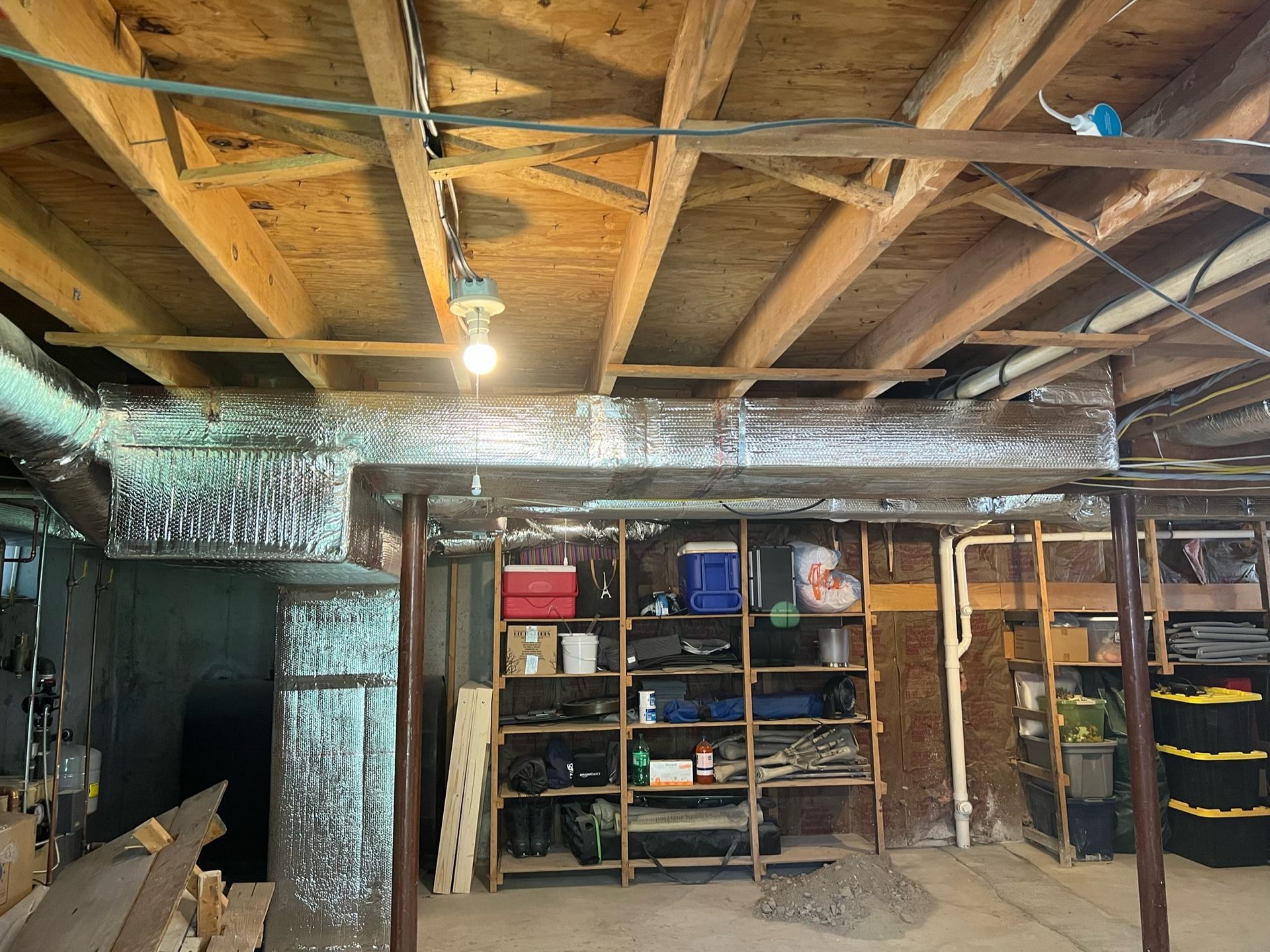 Basement with wooden ceiling, shelving unit, ductwork, and exposed support beams. A bare lightbulb hangs down.