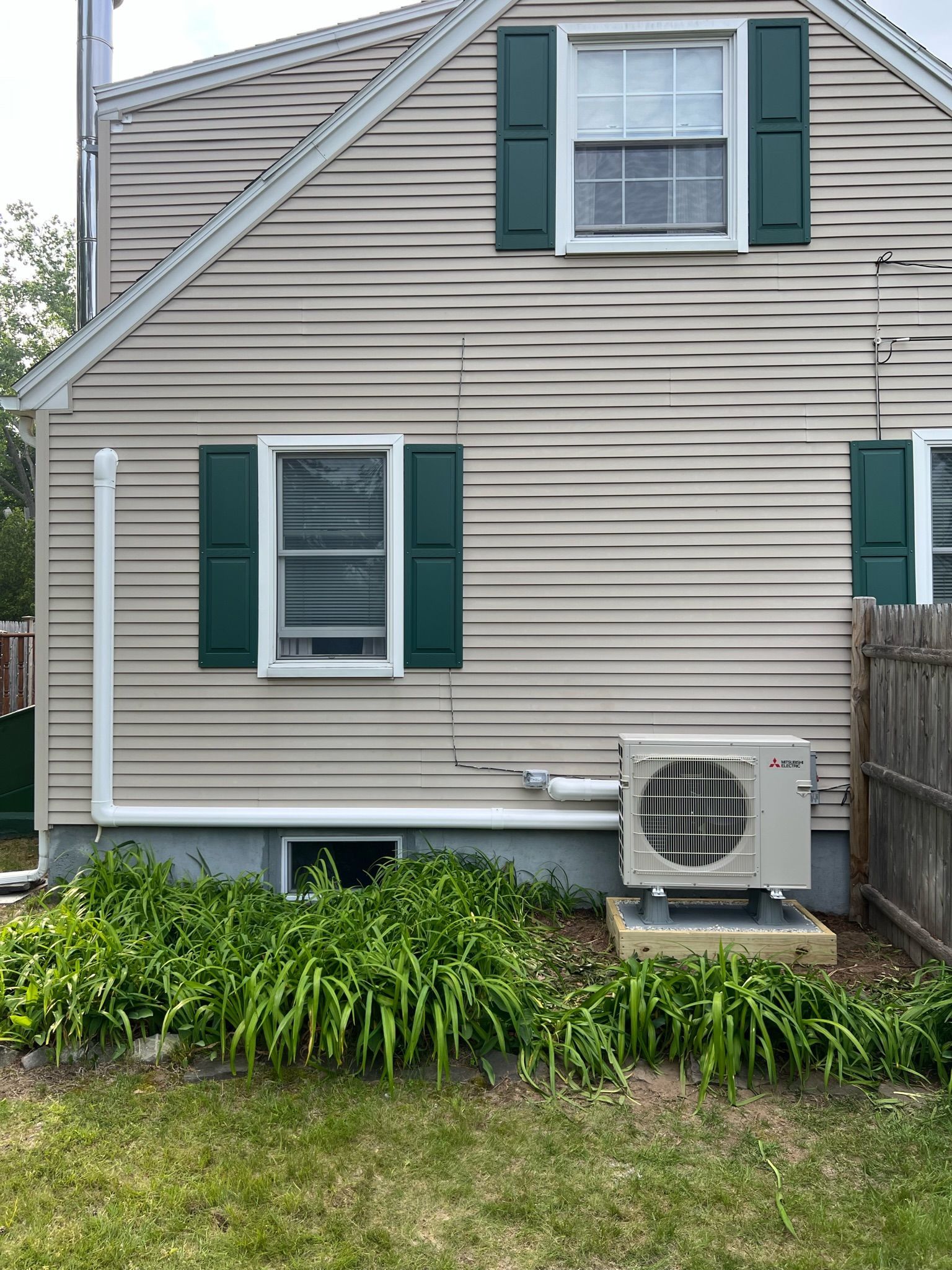 Beige house siding with green shutters, a ground-level AC unit, and overgrown greenery in front.
