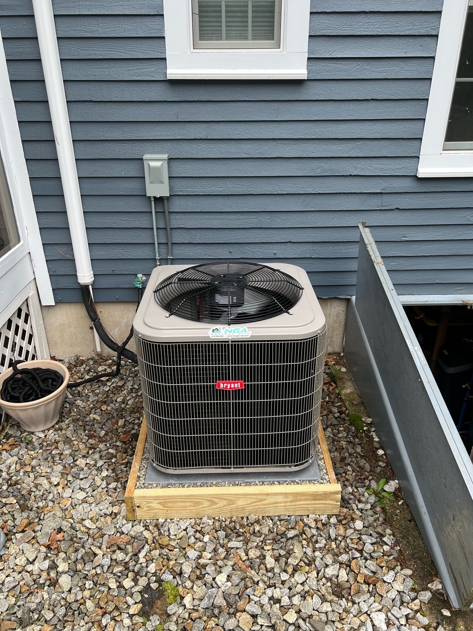 Air conditioner unit on wooden base next to a house with blue siding.