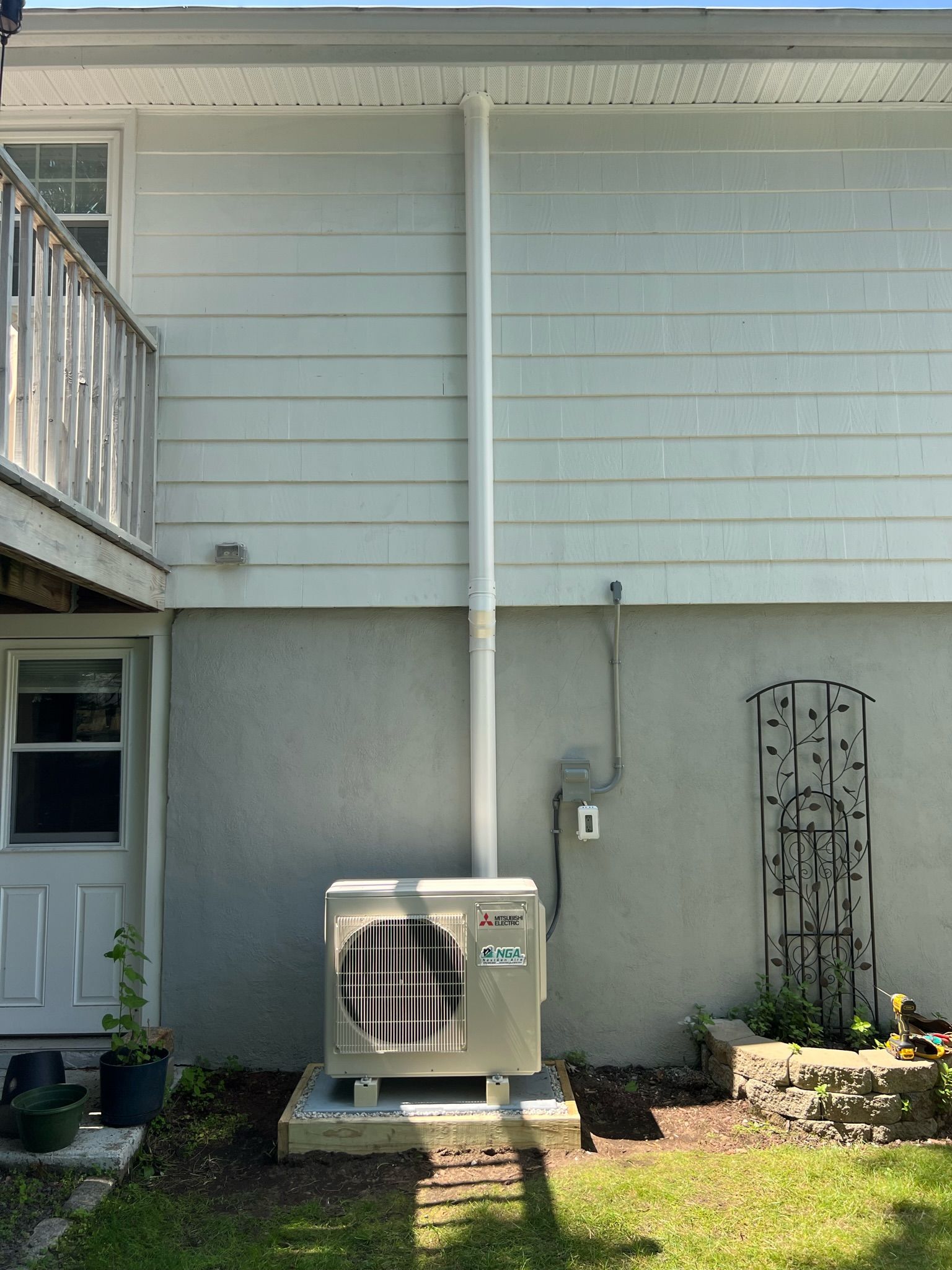 Outdoor air conditioning unit mounted on a concrete pad on the side of a white house with a vertical pipe.