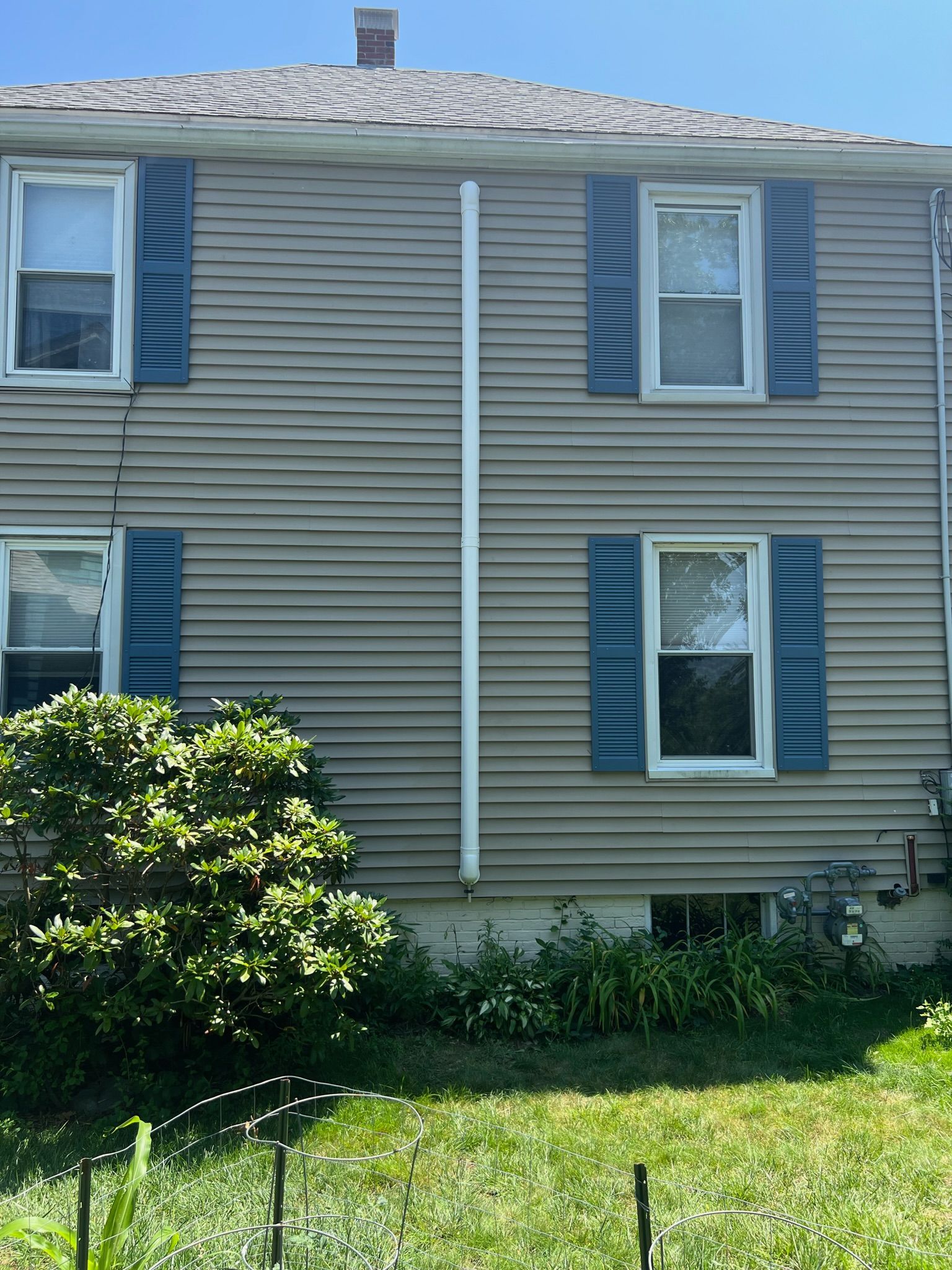 Beige vinyl siding with blue shutters and a white pipe against a sunny day.
