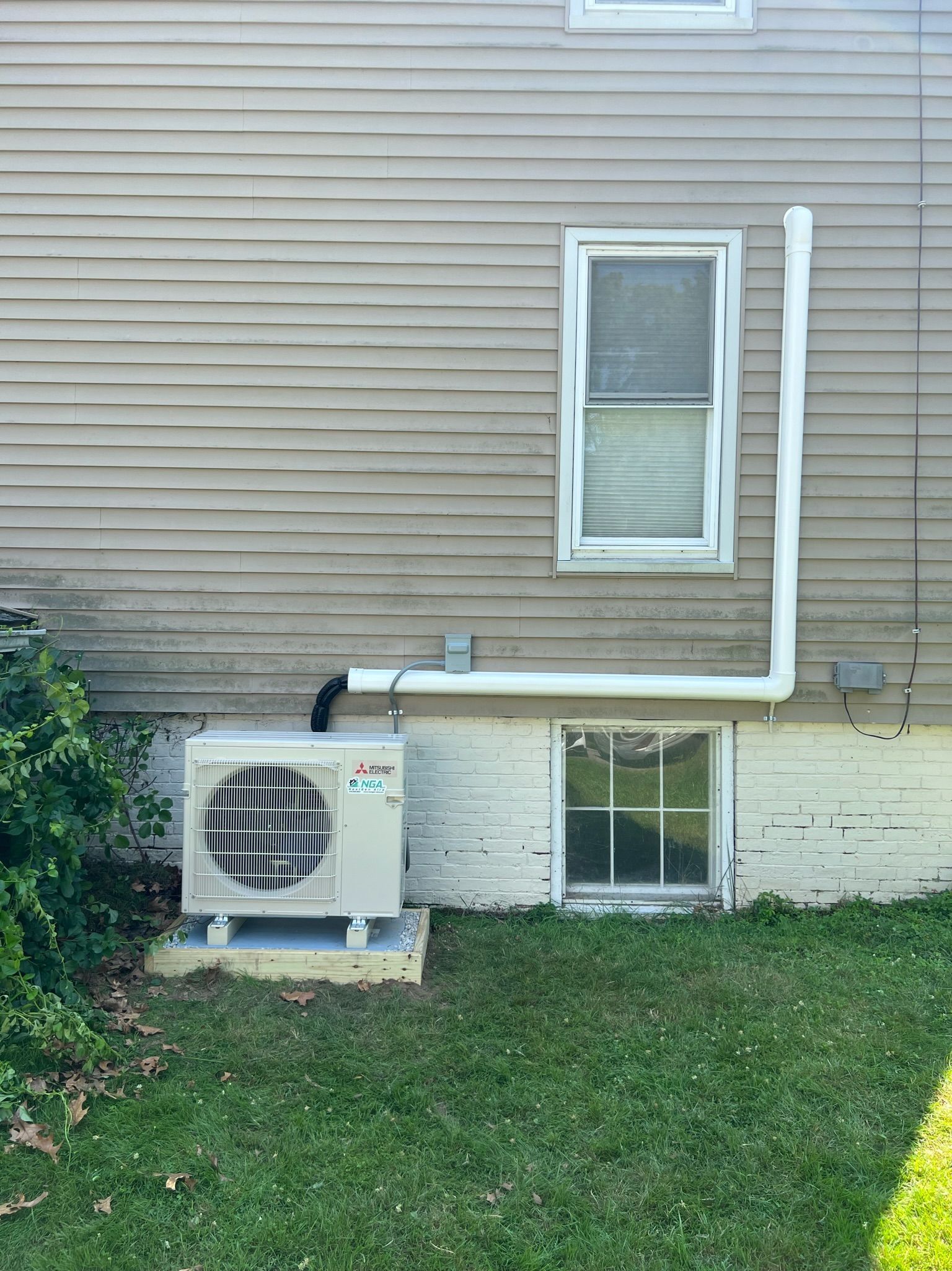 Exterior of a house with an air conditioning unit and white drainage pipe near a window. Green grass and shrubs.